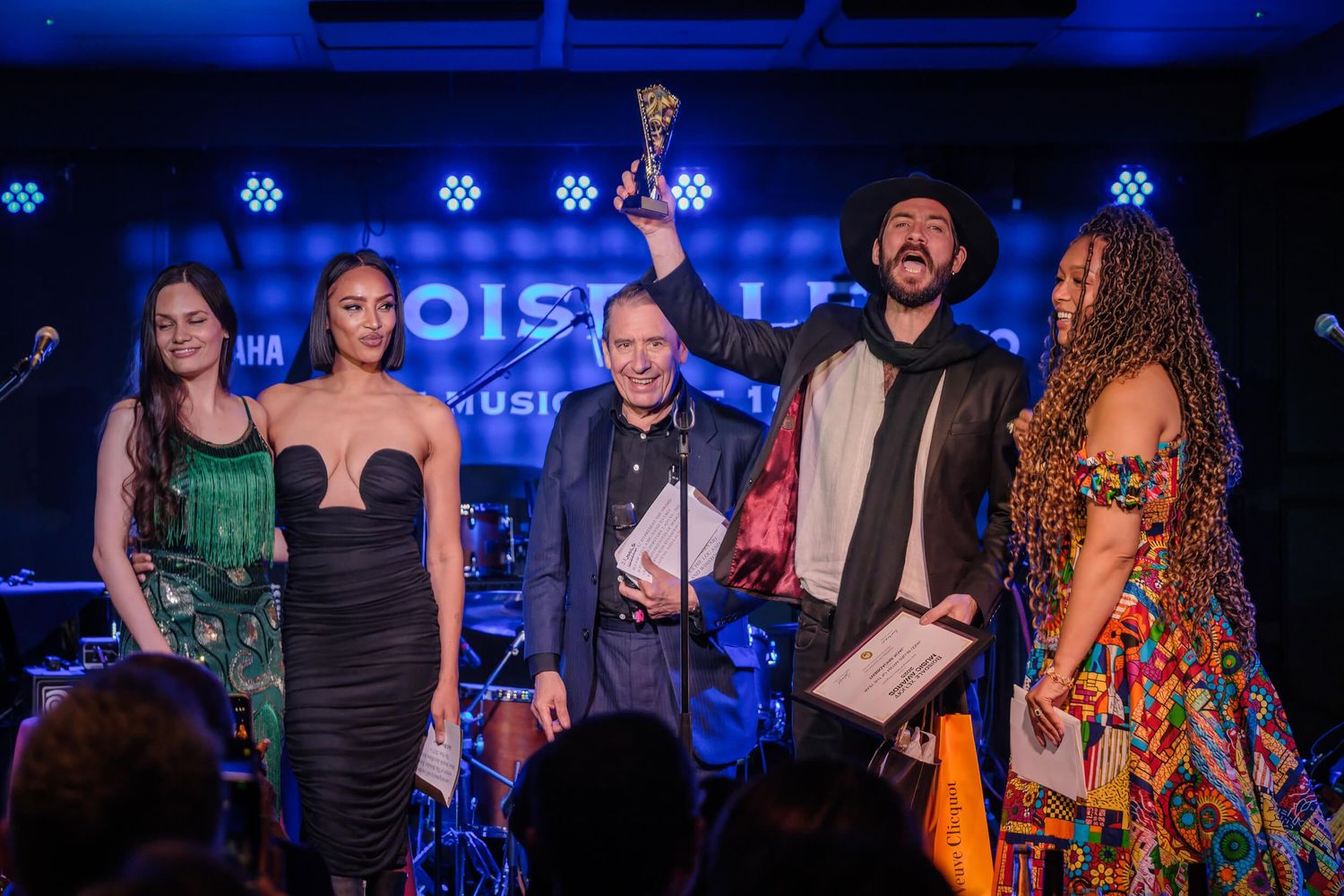 Musicians perform on stage at a concert venue with blue lighting and musical instruments.