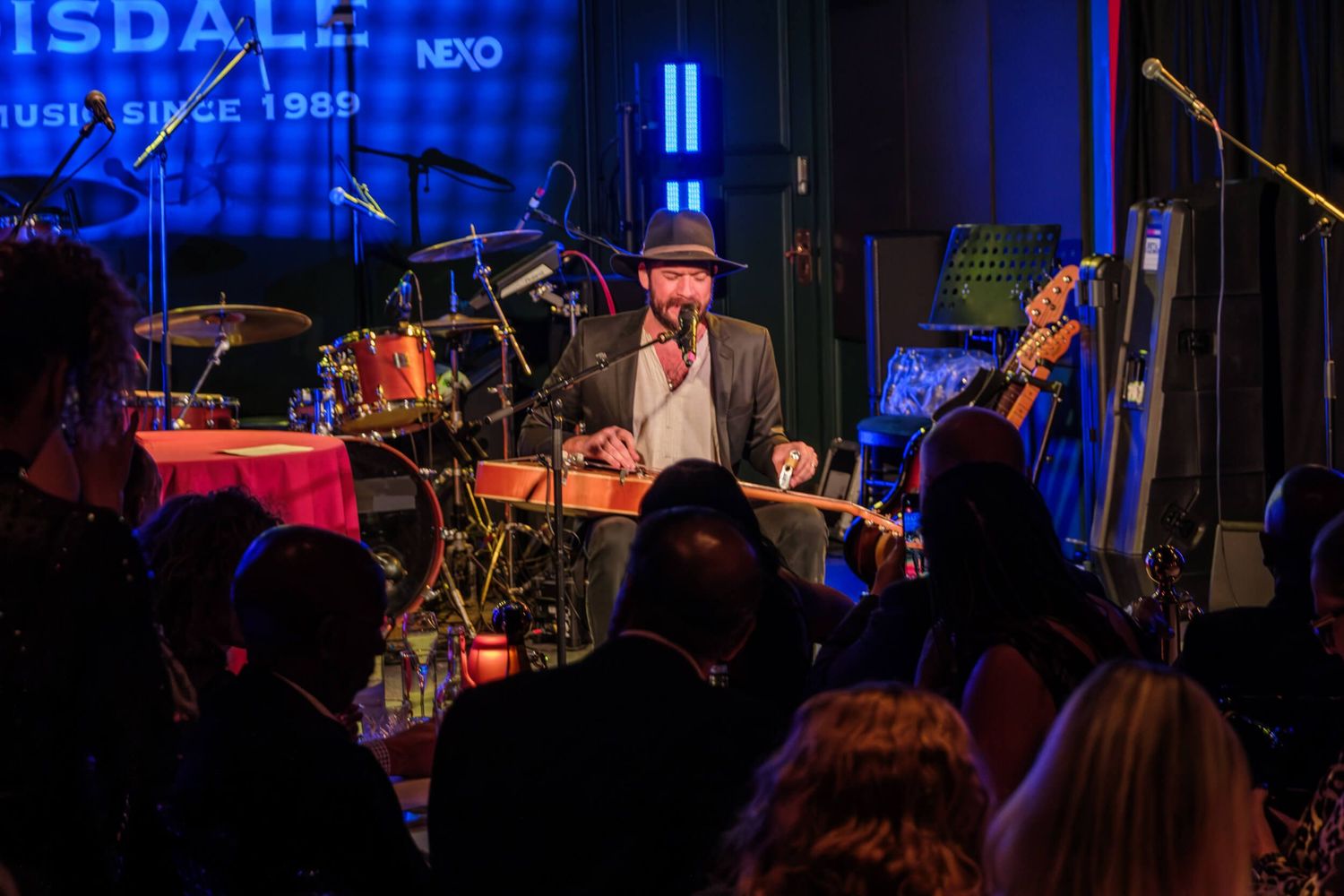 Musicians perform on a dimly lit stage with blue lighting and instruments during a live concert performance.