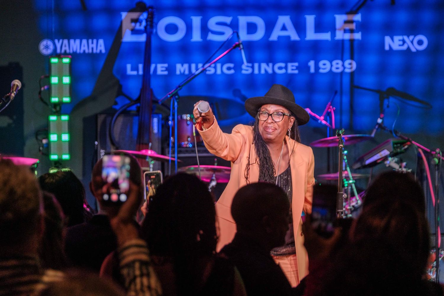 A performer in a brown hat sings on stage with colorful blue and purple lighting in the background.