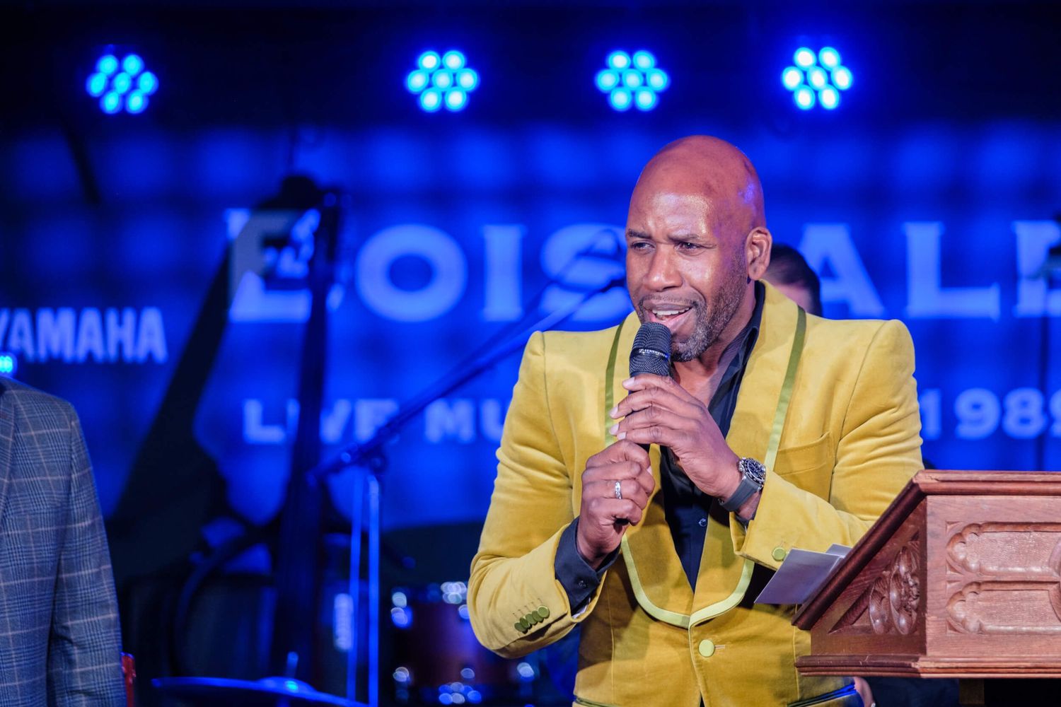 Performer in yellow jacket sings into microphone with blue lighting effects illuminating the stage.