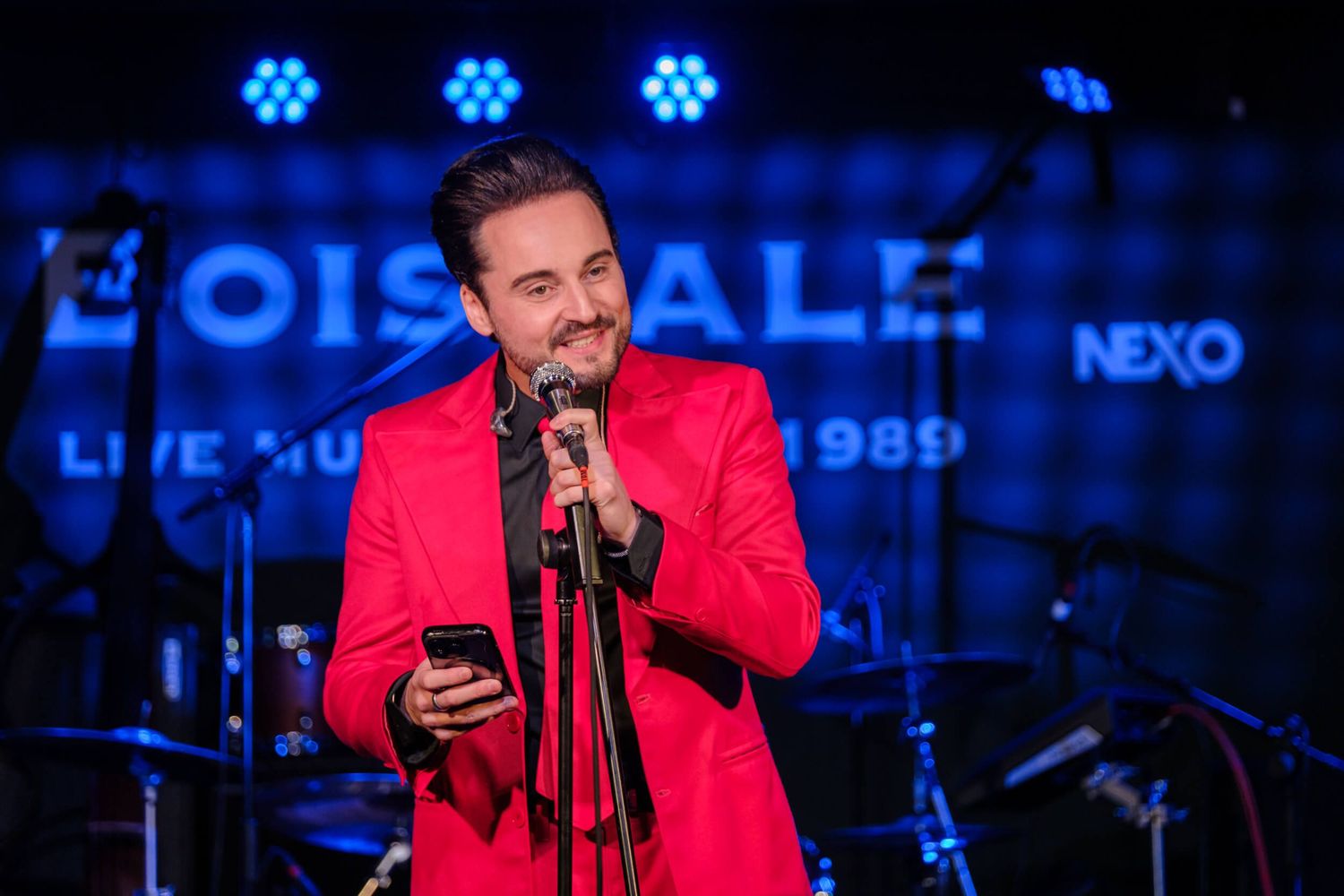 A performer in a bright red jacket sings into a microphone while looking at a phone on stage with blue lighting.