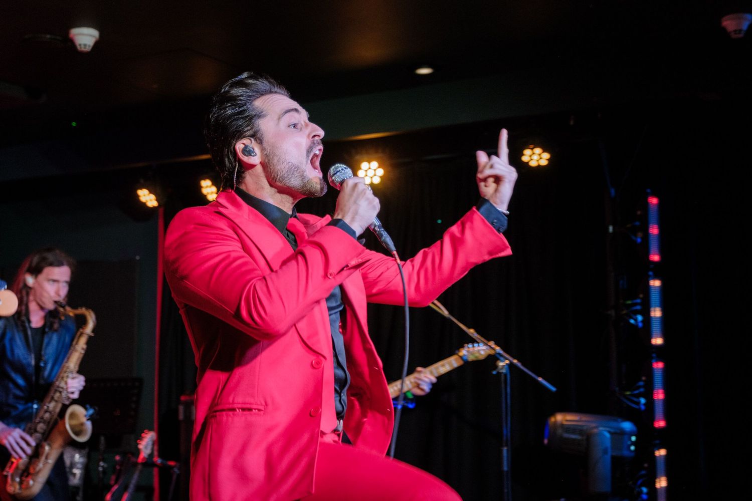 A performer in a bright red jacket sings passionately on stage during a live concert performance.