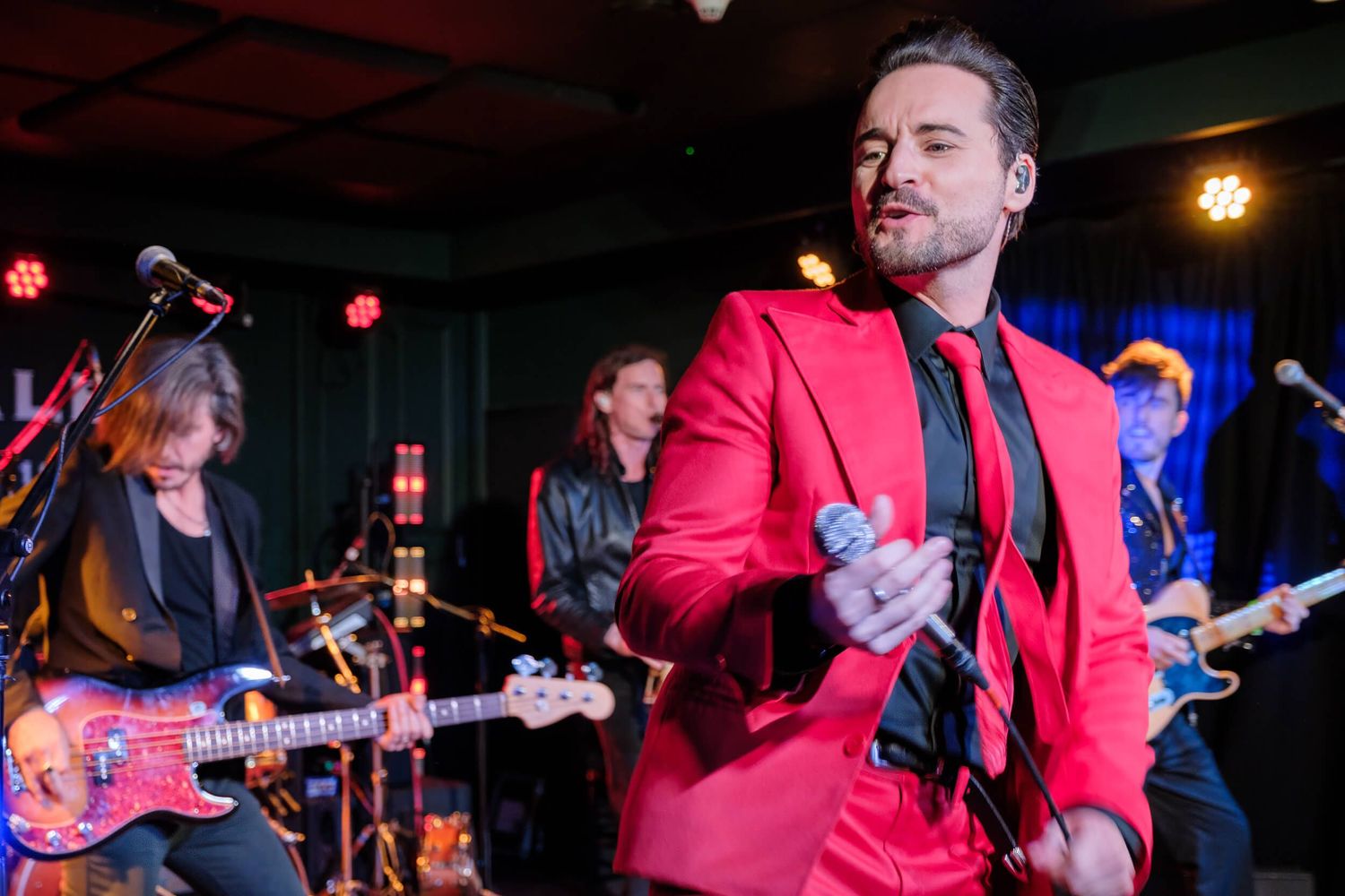 Musicians in red shirts perform on stage with guitars and instruments under colorful stage lighting.