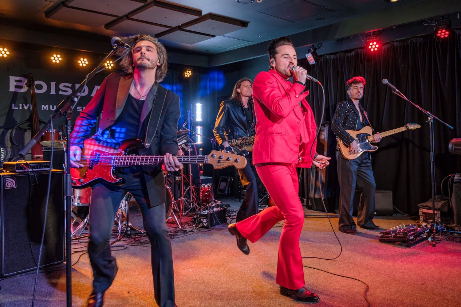 Musicians perform energetically on stage in red outfits under colorful stage lighting at a live concert venue.
