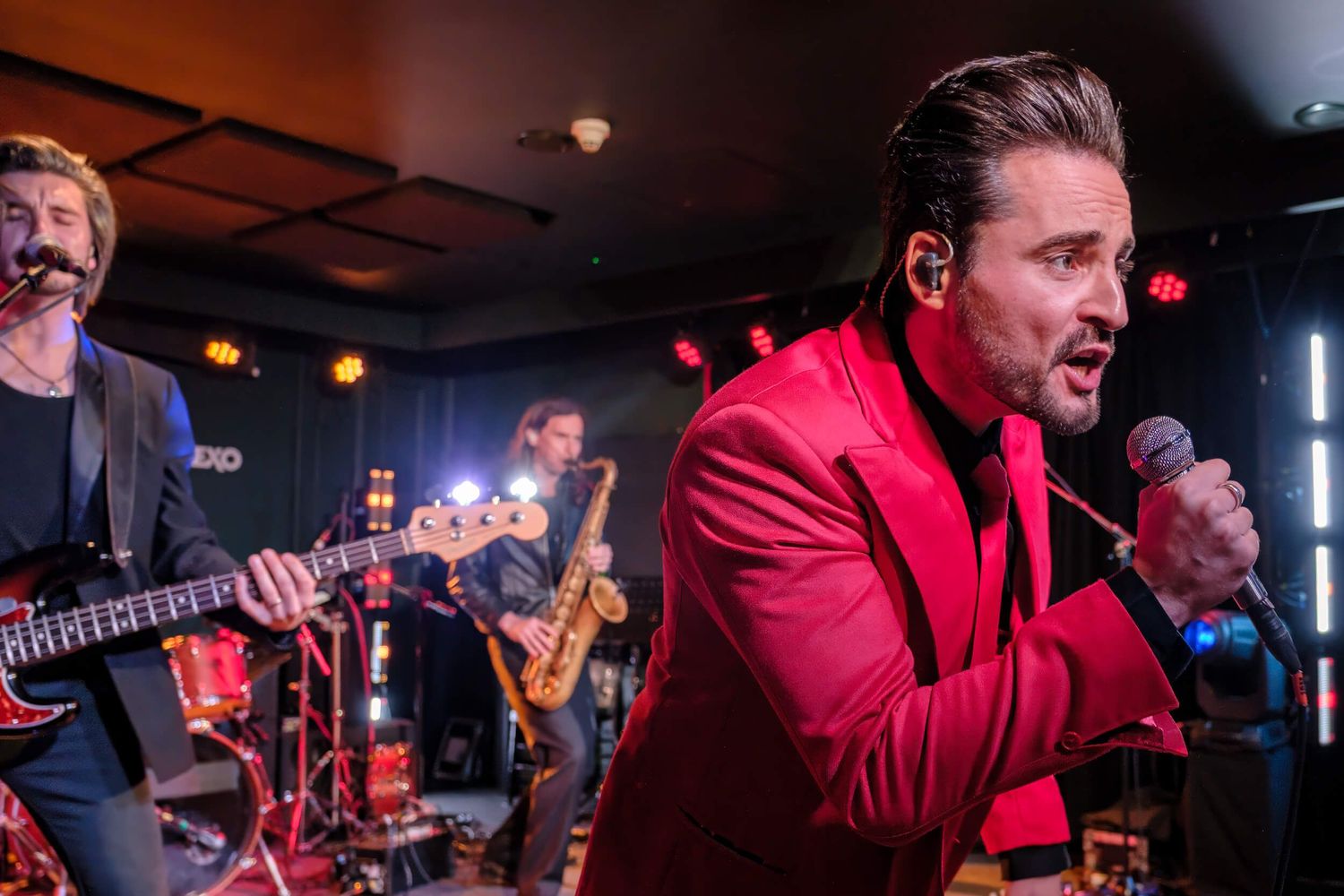 Live band performs on stage with singer in red jacket and musicians playing instruments at dimly lit venue.