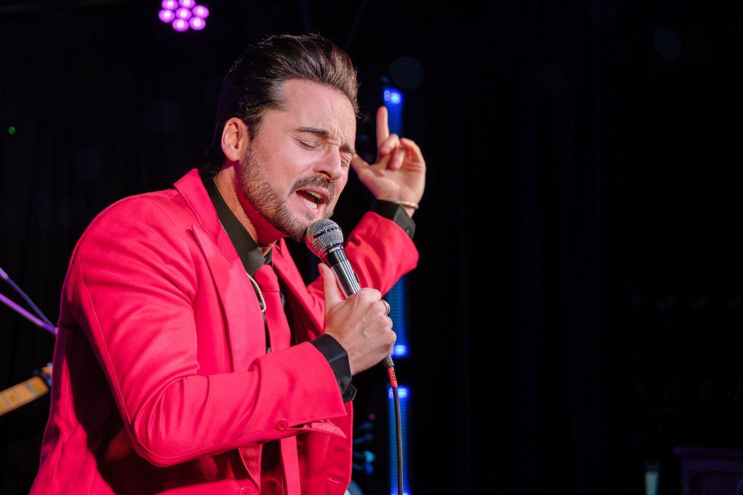 A performer in a bright red jacket sings passionately on stage during a live concert performance.