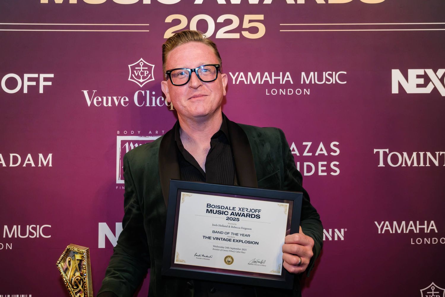 Someone in glasses holds up an award certificate while smiling at a formal event with branded sponsor backdrop.