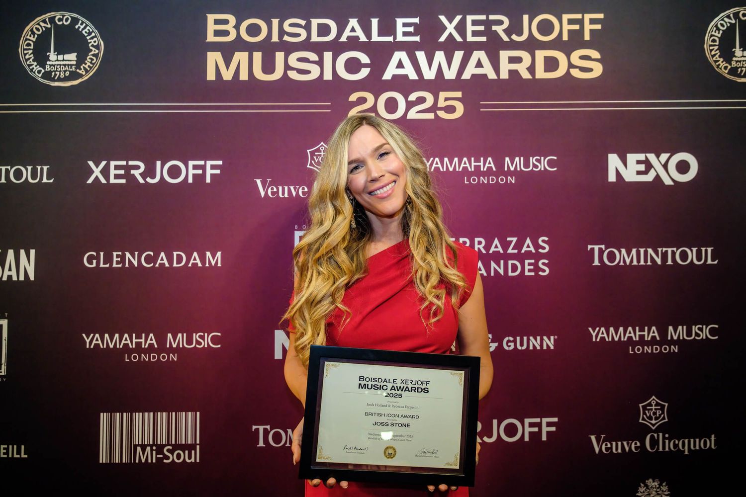 A person in a red dress holds an award at a music awards ceremony with sponsor logos visible in the background.
