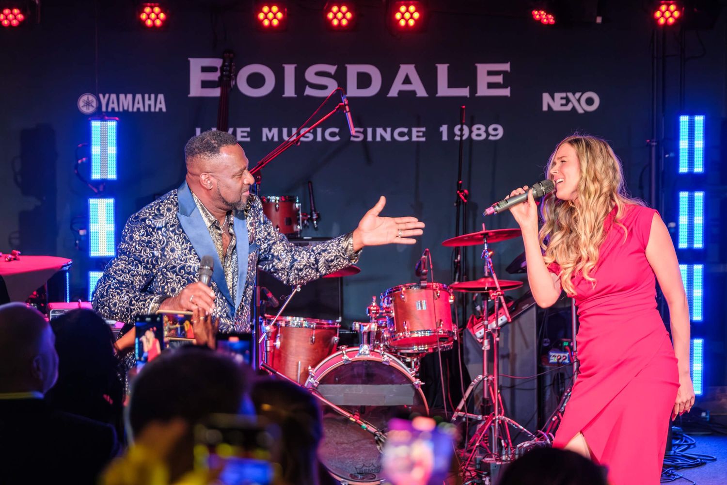 Performers on stage during a concert with red lighting and musical equipment visible in the background.