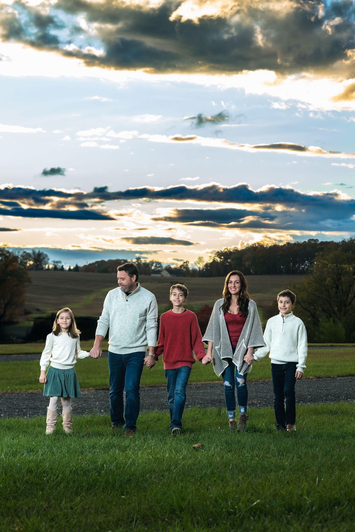 A family walks together across a grassy field at sunset with dramatic clouds in the sky above.