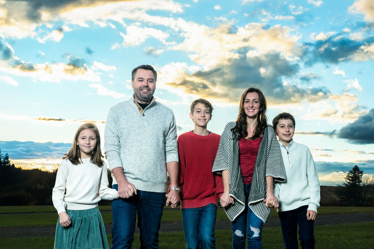 A family in coordinated red and white outfits poses together outdoors during sunset against a dramatic cloudy sky.