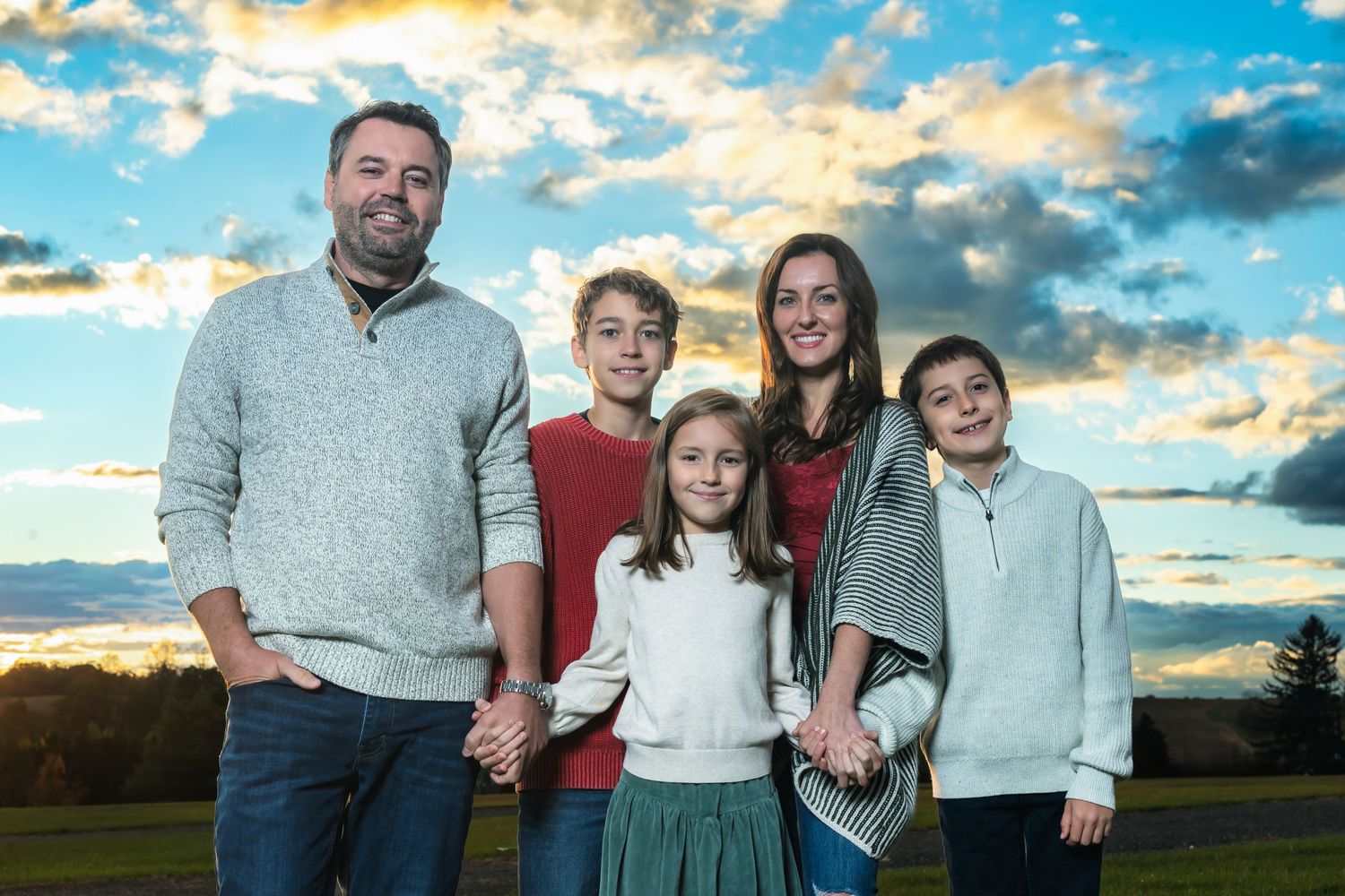 Family poses together in front of dramatic sunset sky with clouds in outdoor fall setting.
