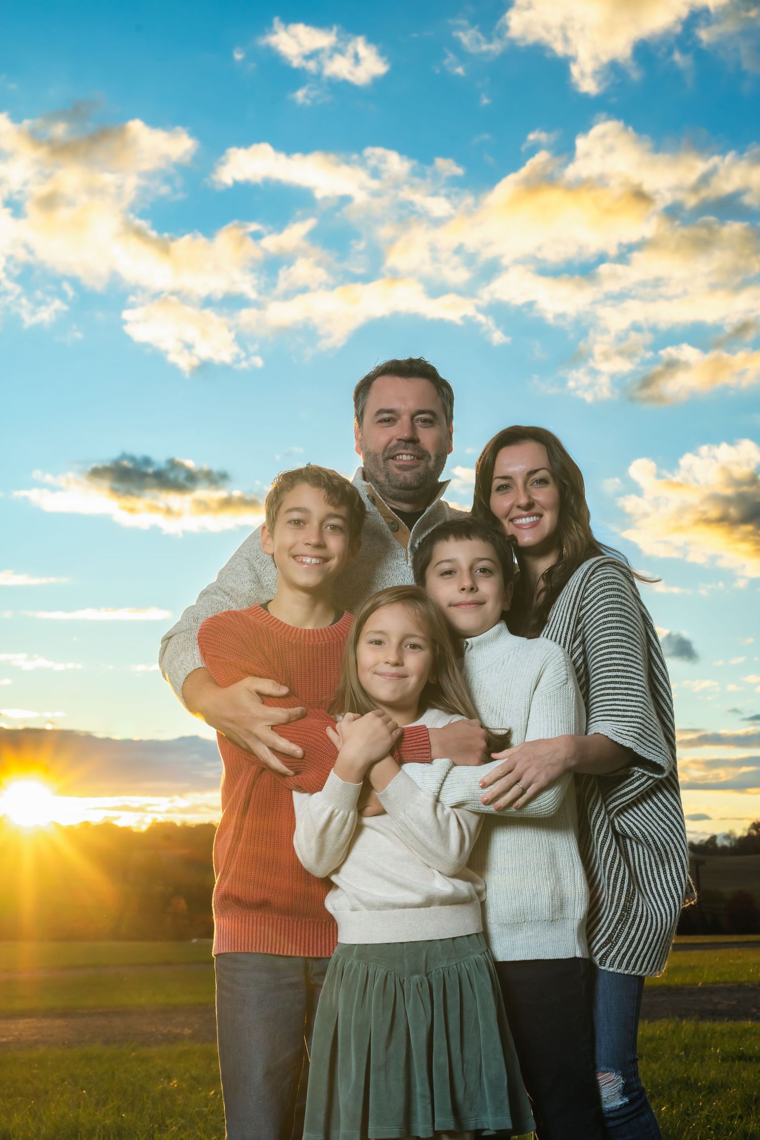 A family poses together outdoors at sunset with a dramatic sky and golden sunlight in the background.