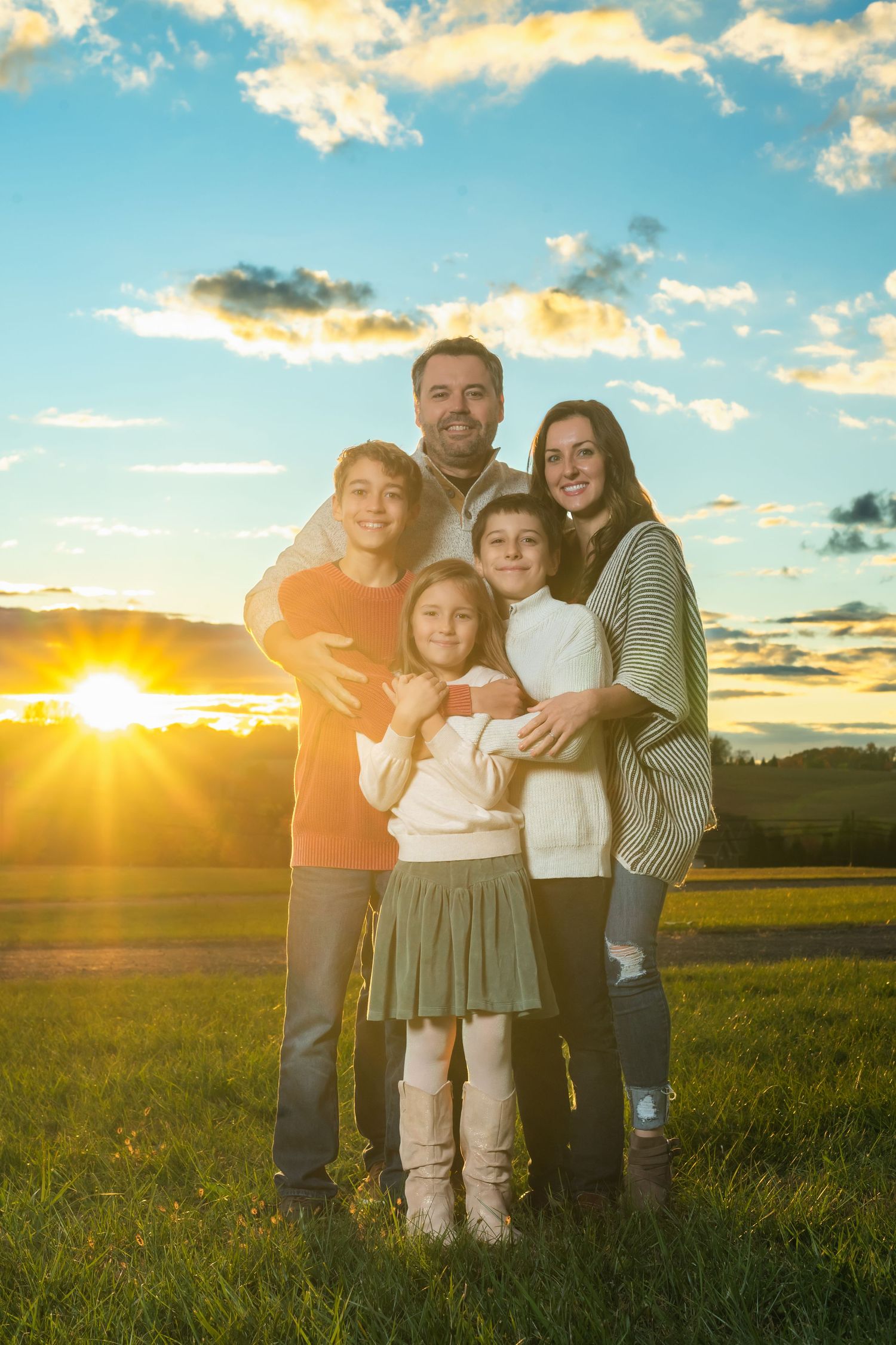 A family poses together in a grassy field during golden hour sunset with dramatic clouds in the sky.