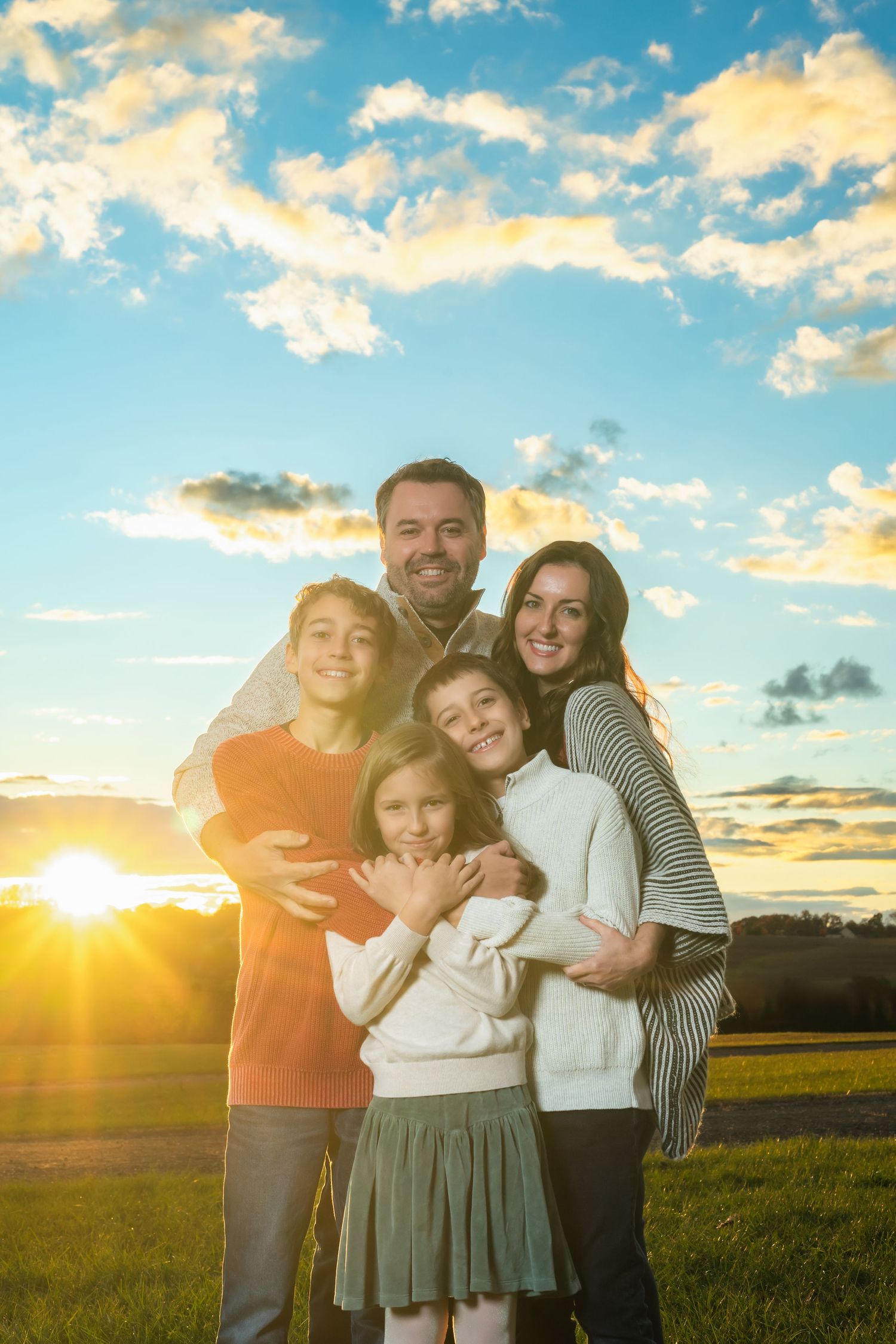 A family embraces while standing in a grassy field at sunset with golden sunlight and clouds in the background.