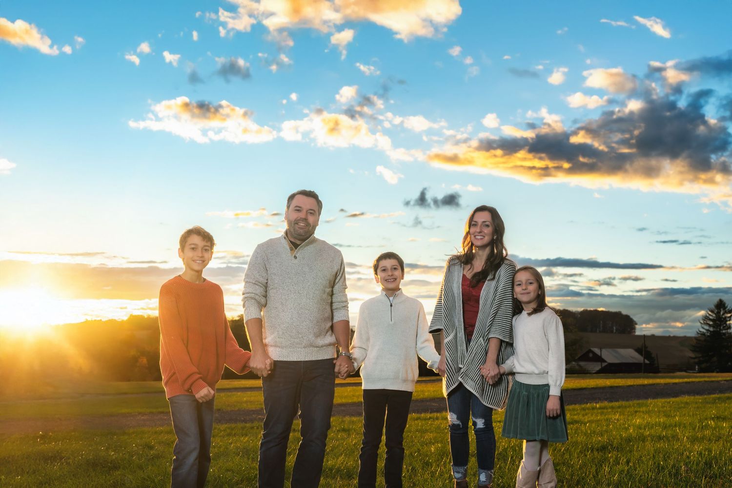 A family stands together in a golden sunlit field during sunset with dramatic clouds in the background.