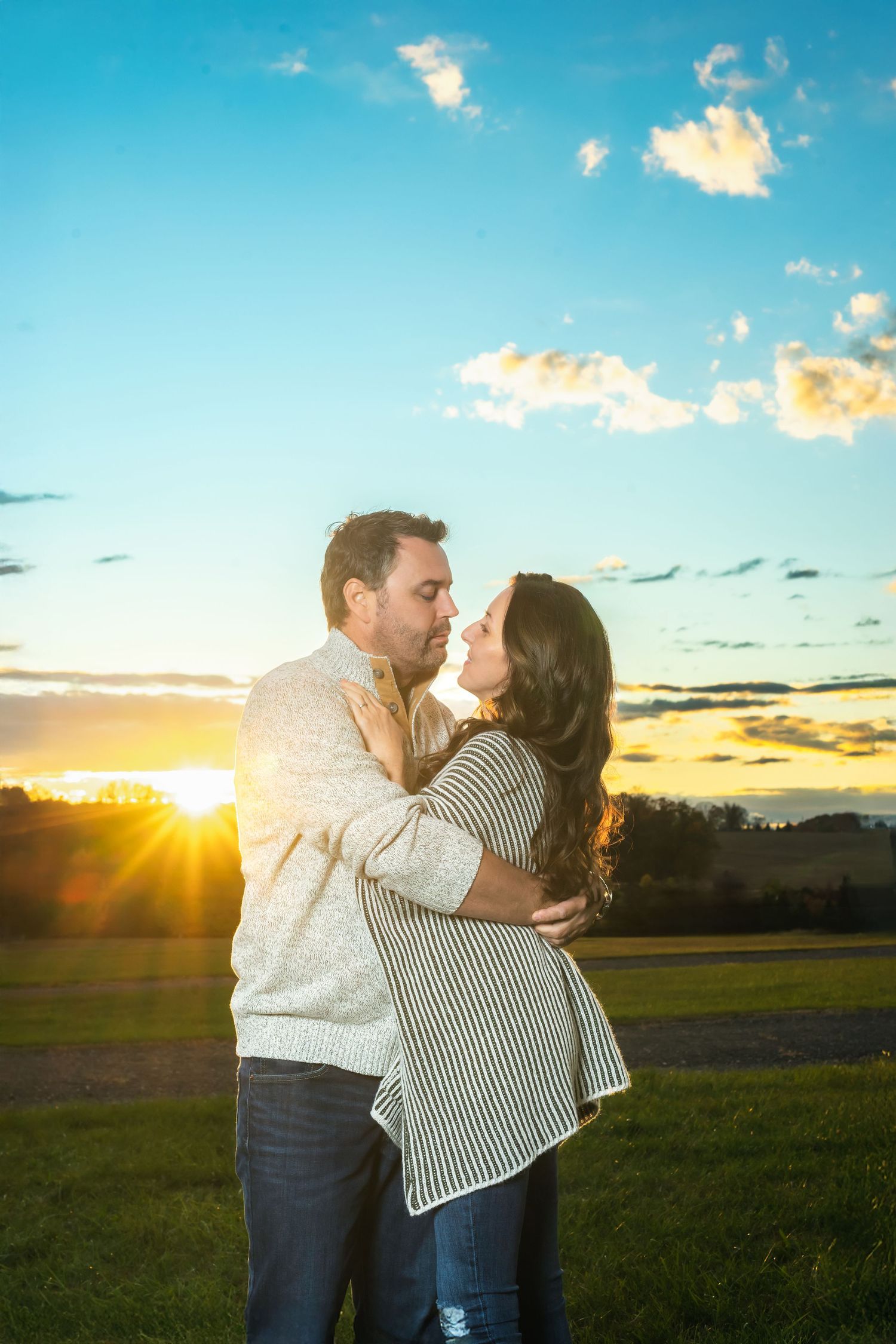 A romantic couple embraces in a field at sunset with golden clouds and a vibrant blue sky in the background.