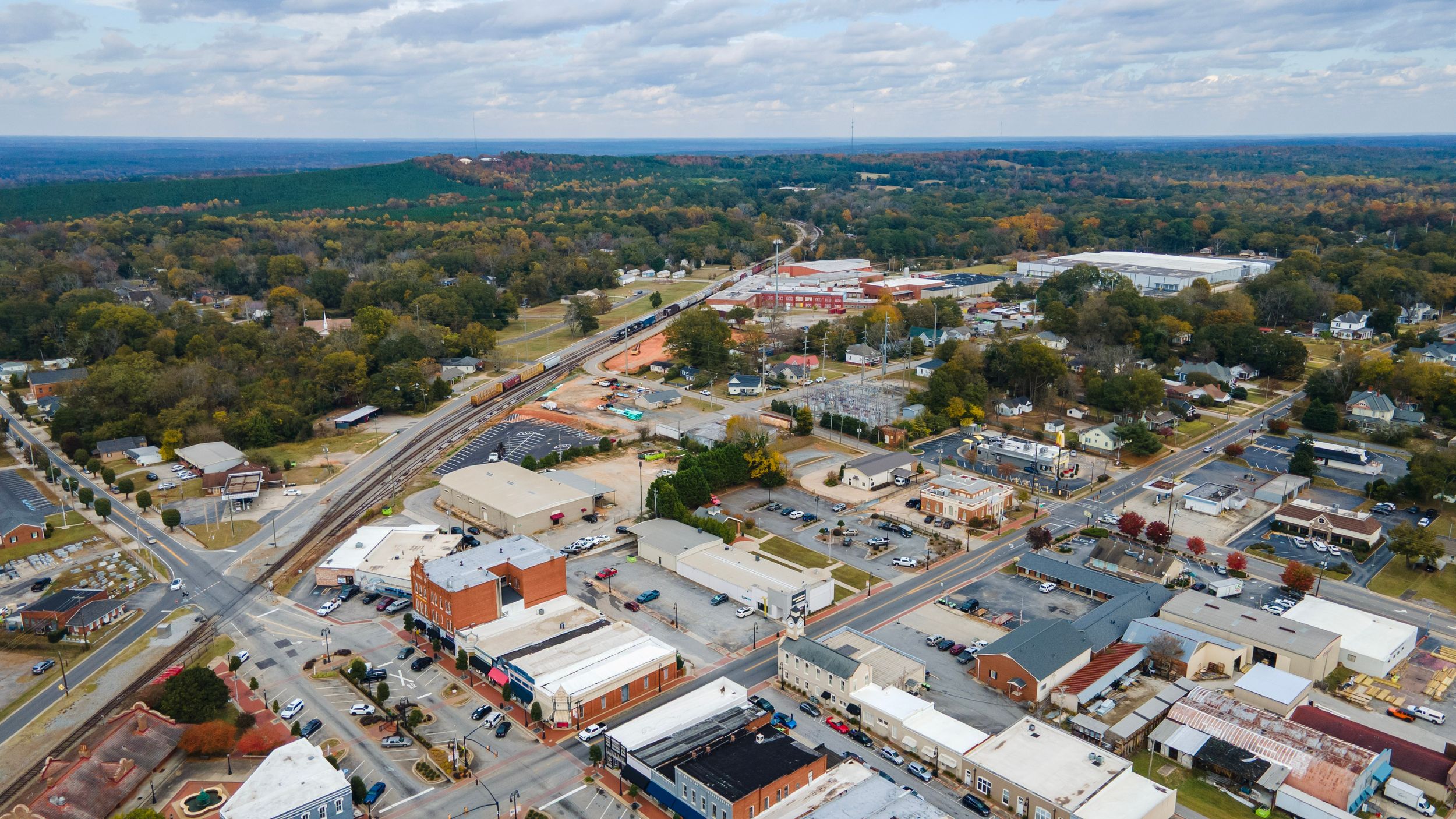 Aerial panoramic view of a small town with buildings and roads surrounded by green farmland under a cloudy sky.