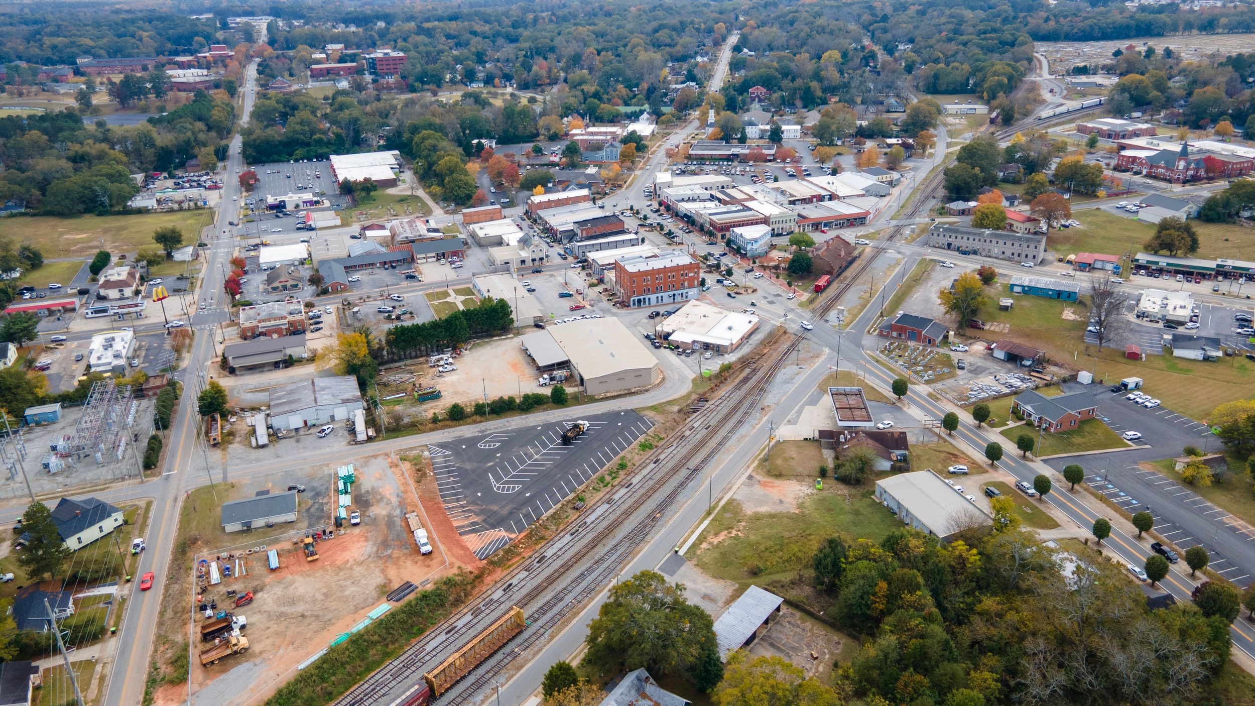 Aerial panoramic view of a small town with railroad tracks, buildings, and autumn trees along a winding road.