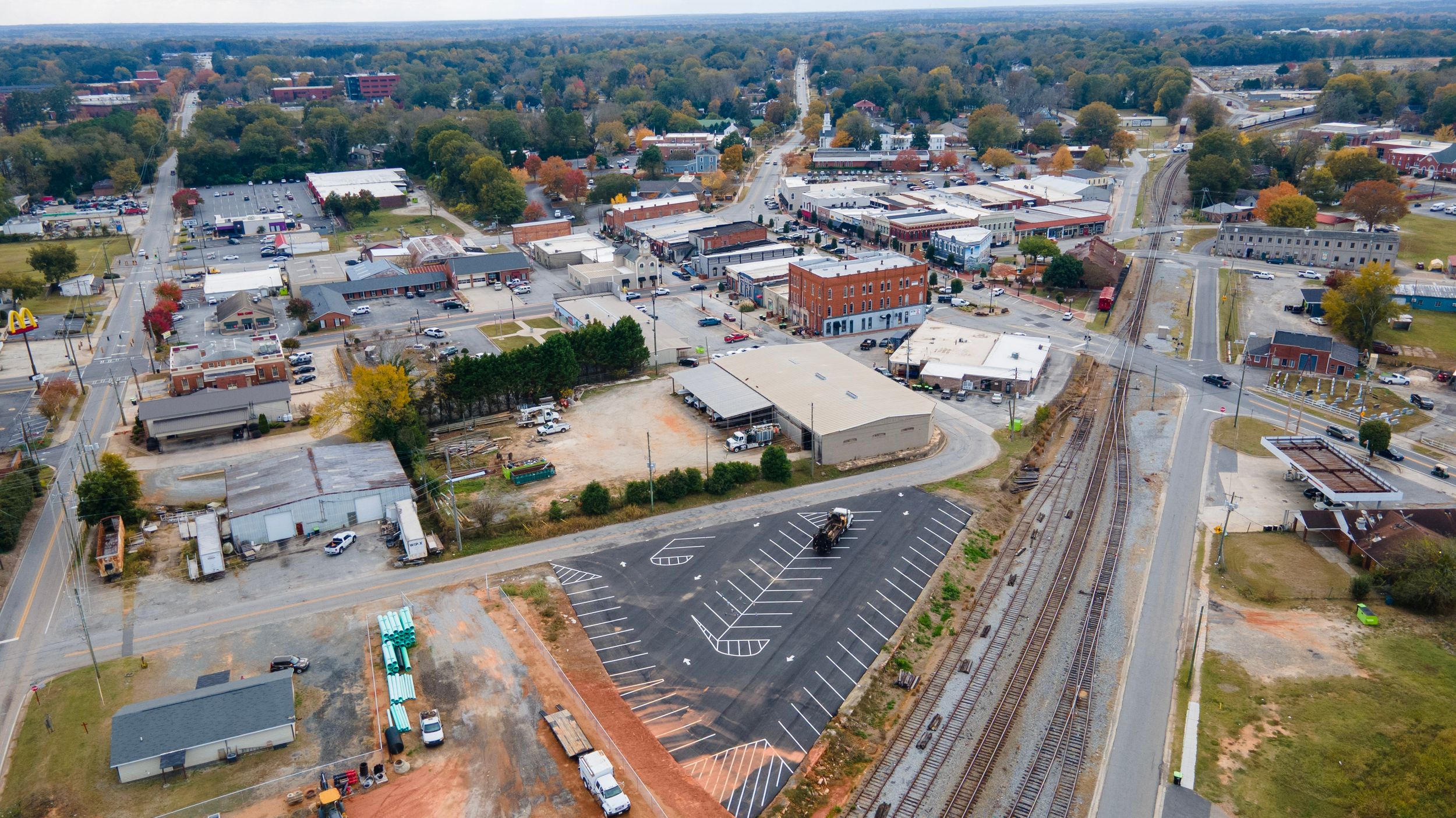 Aerial panoramic view of a small town with commercial buildings, parking lots, and tree-lined streets on a sunny day.