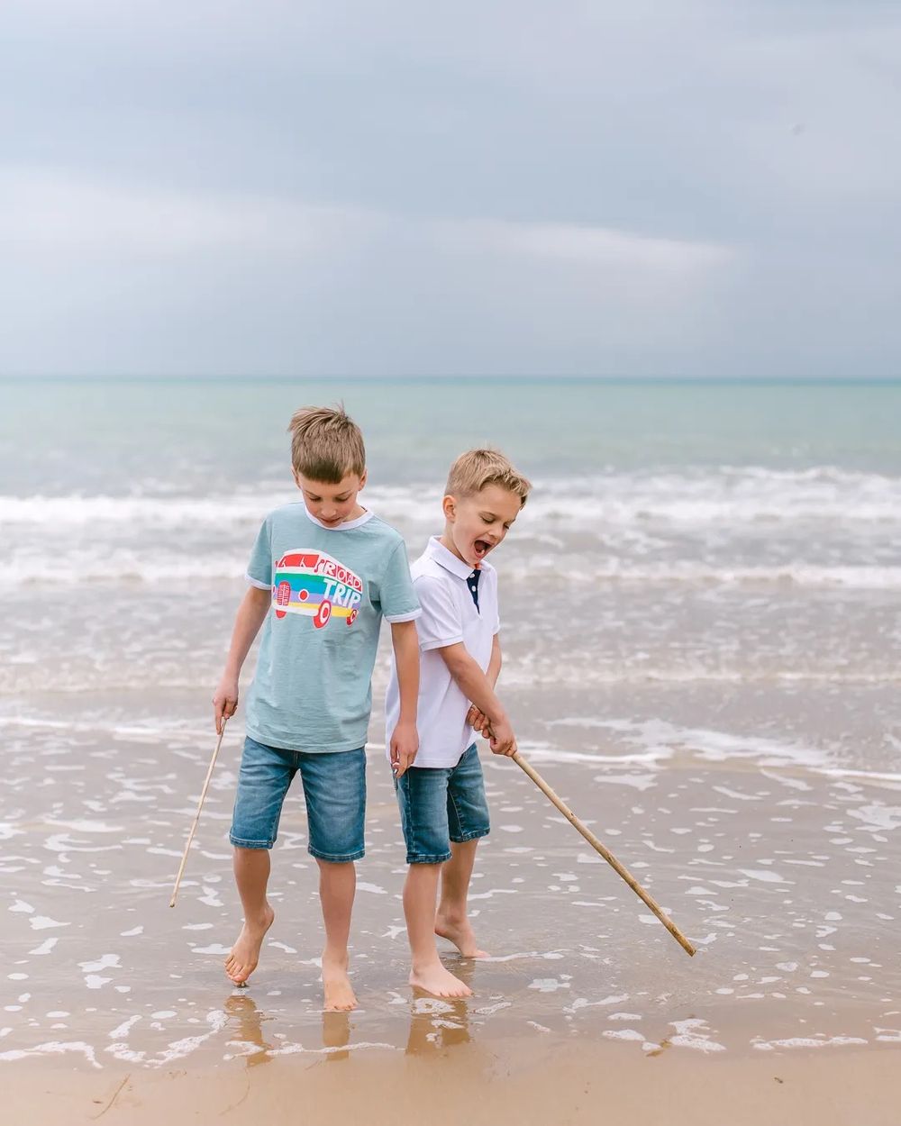 Two boys standing in shallow ocean waves, smiling while playing at the beach.
