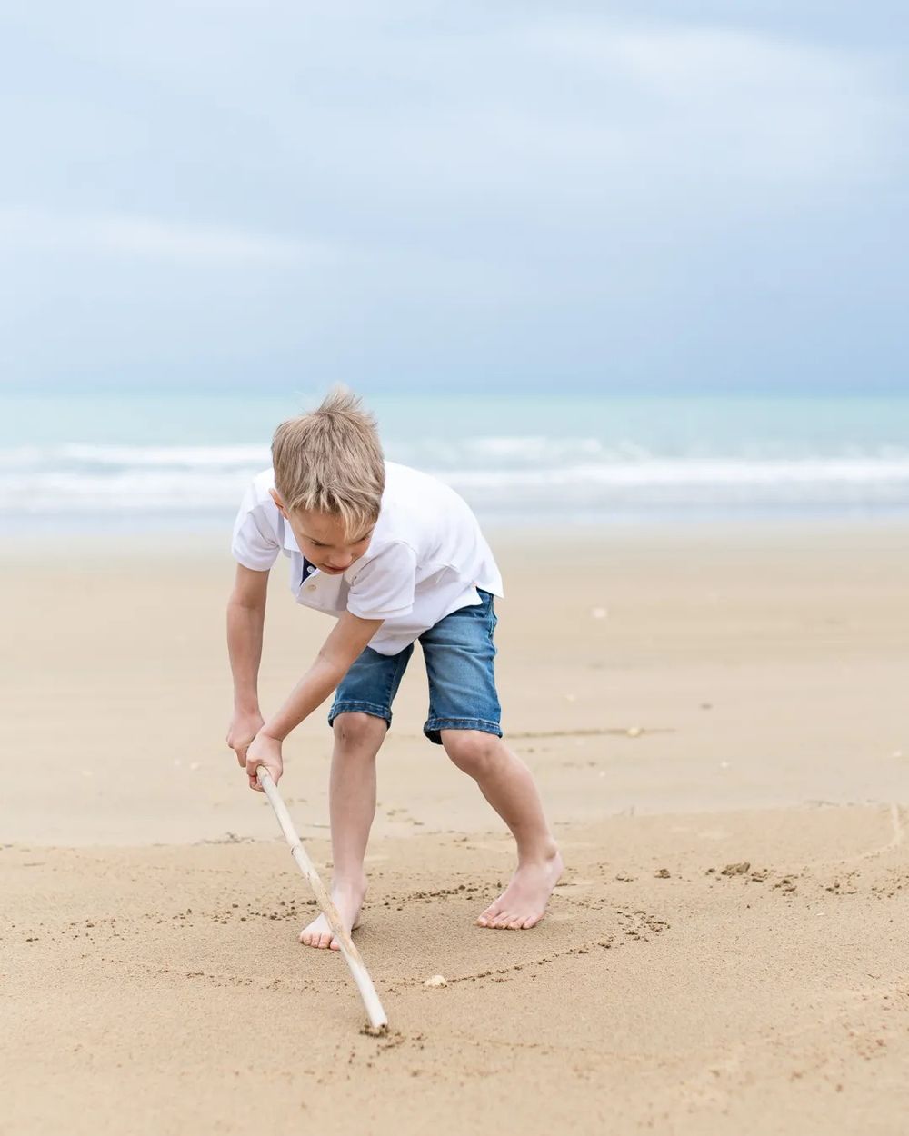 Young boy digging in wet sand by the ocean on a cloudy beach day.
