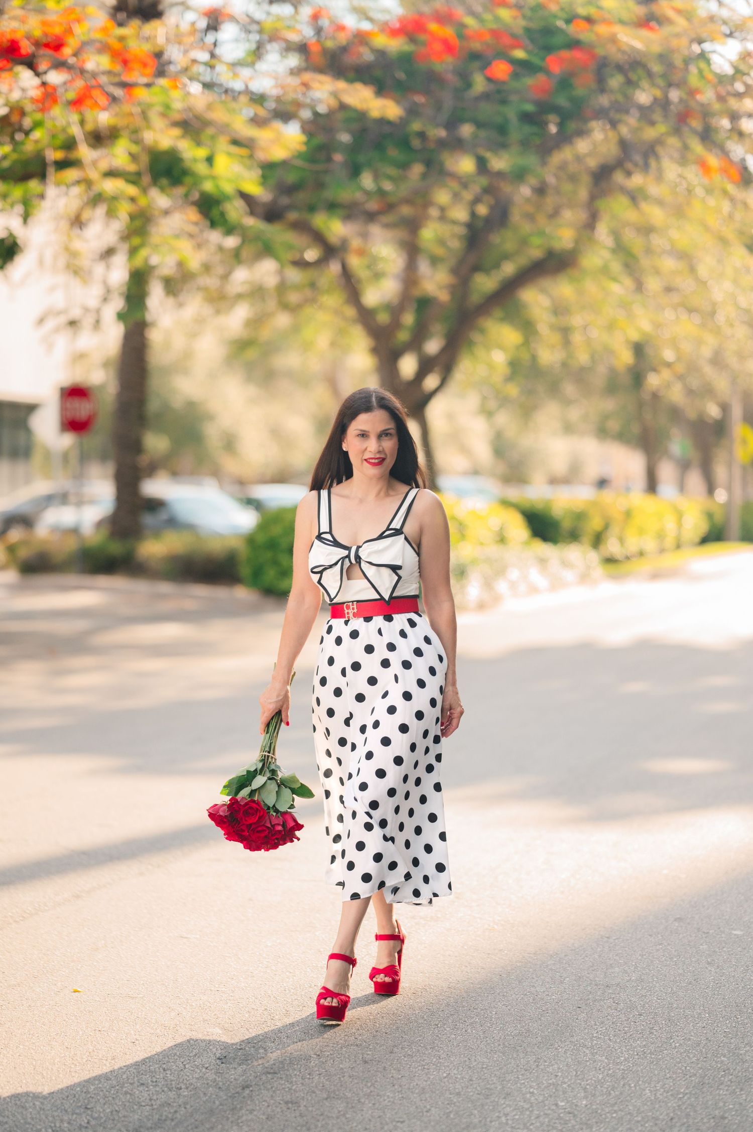 A woman in a polka dot dress poses outdoors with red roses during autumn with colorful fall foliage in the background.