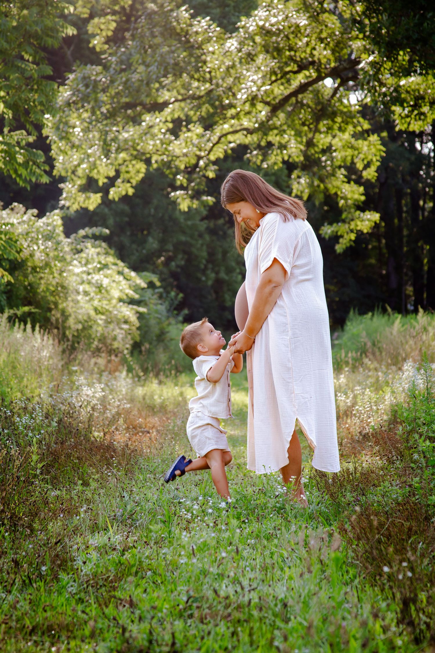 Mother and child playing together in sunlit meadow wearing white summer outfits.