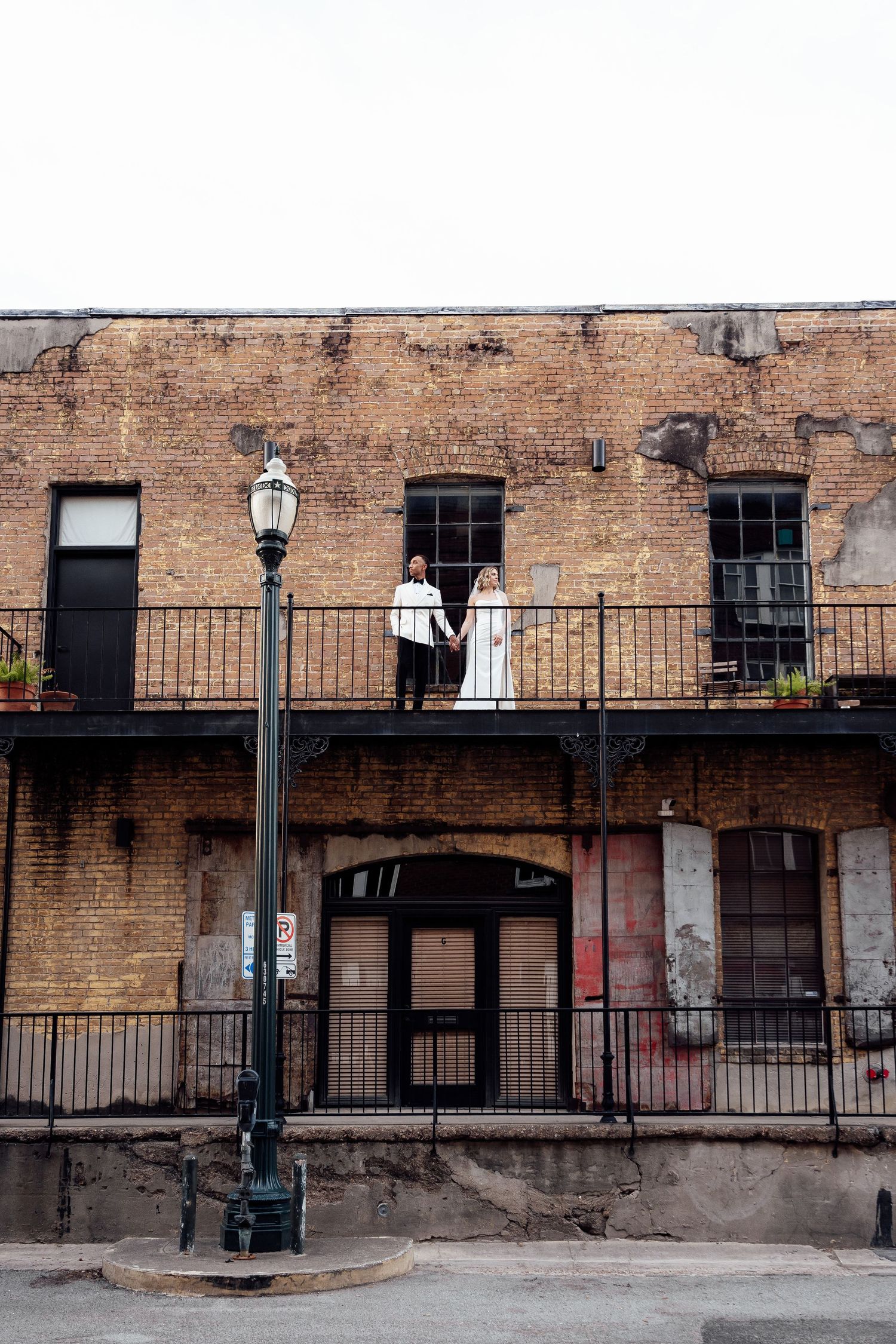 People in white wedding attire stand along a rustic brick wall with iron railing at a historic venue.