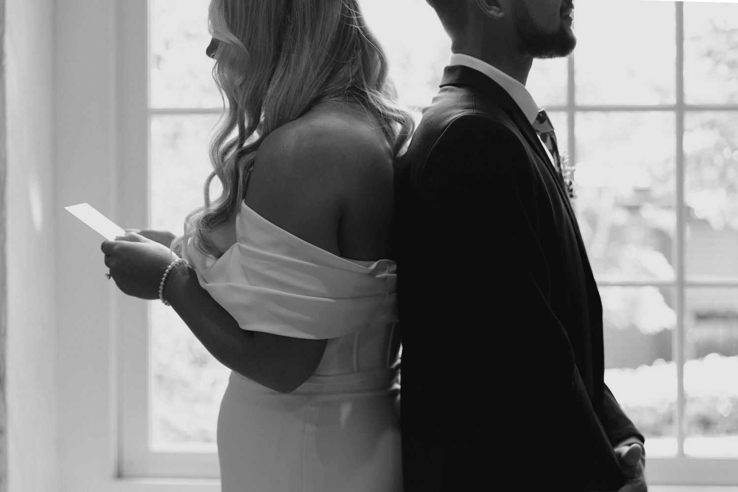 A black and white photo showing a couple standing back to back looking at their phones in formal attire.