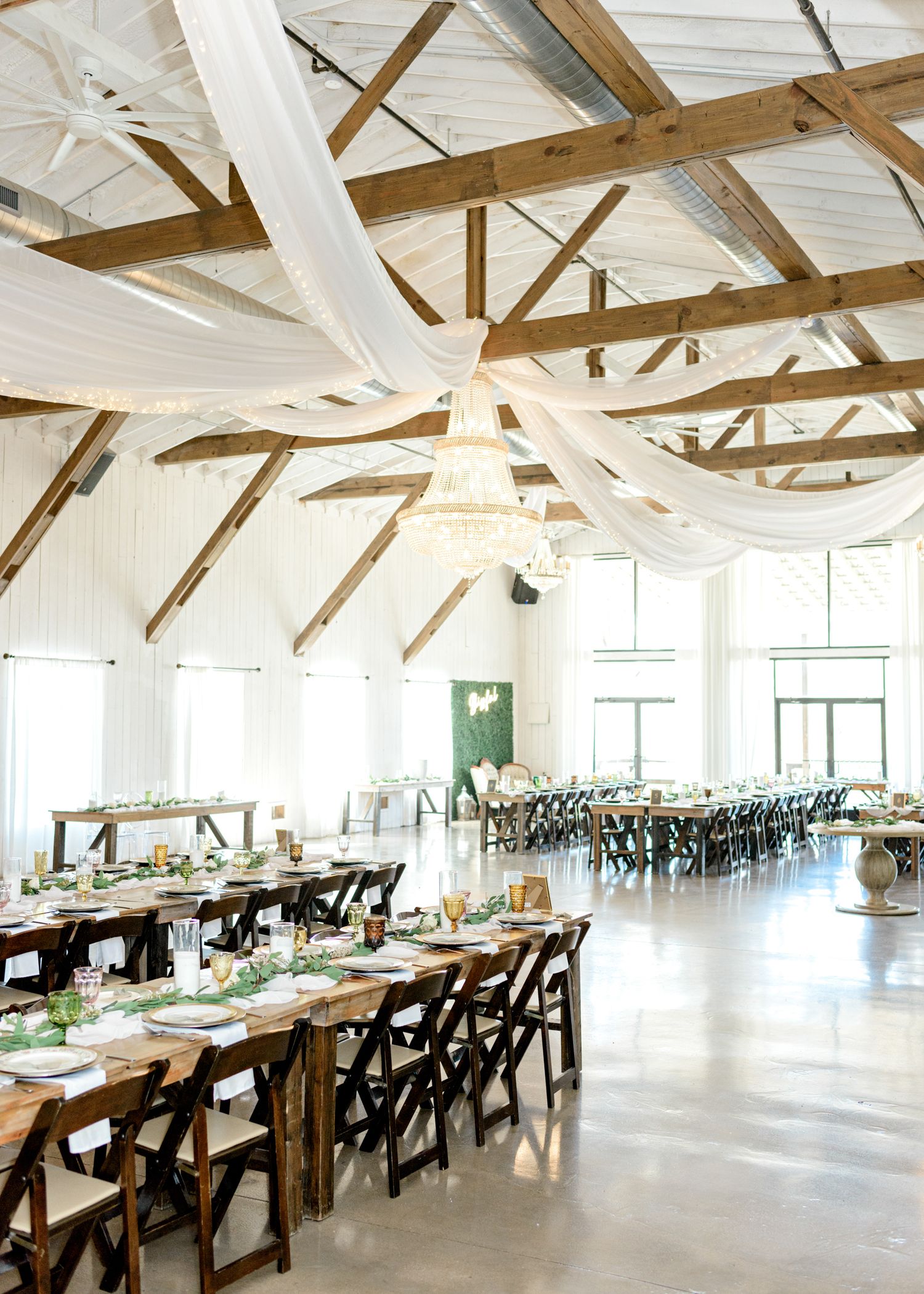 Elegant reception hall with exposed wooden beams decorated with flowing white fabric and rustic dining tables.