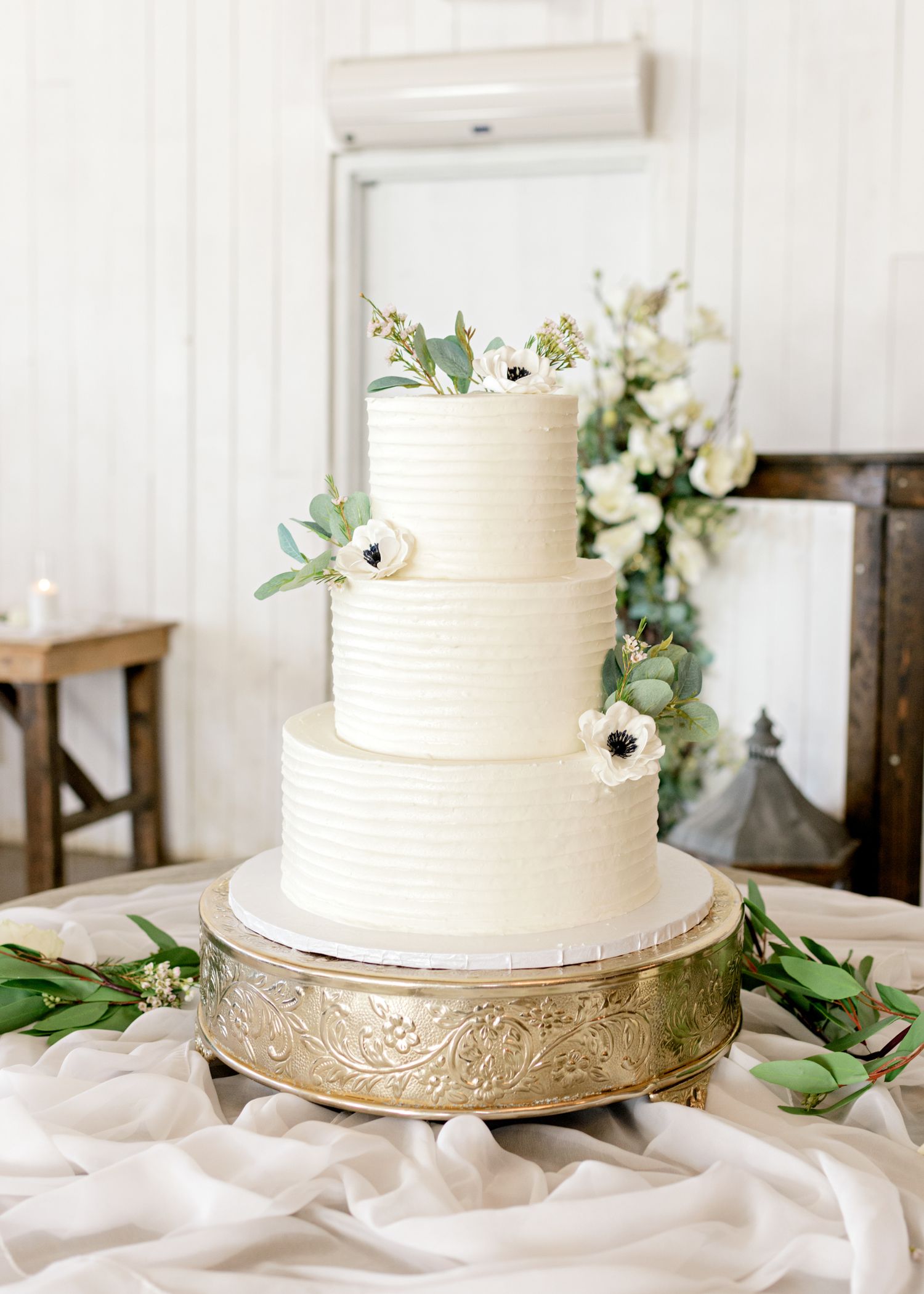 Three-tiered white wedding cake decorated with anemone flowers and eucalyptus on a gold stand.