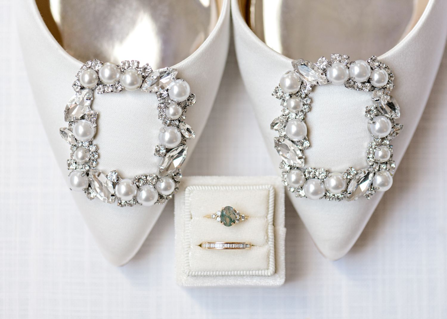 White satin bridal shoes with ornate crystal buckles next to an engagement ring box.
