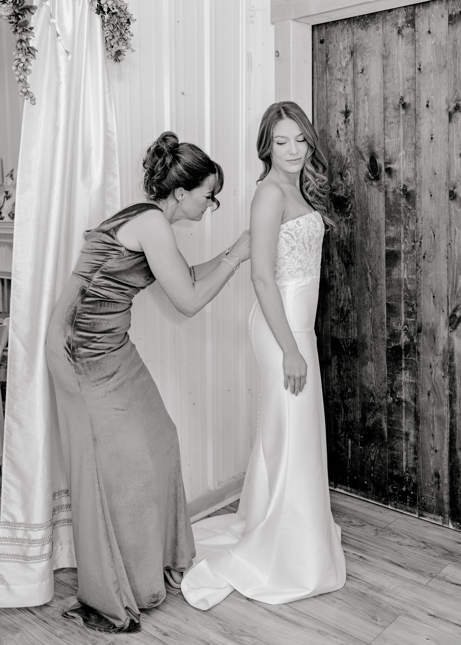 An elegant bridal preparation moment showing final dress adjustments against a rustic wooden backdrop.