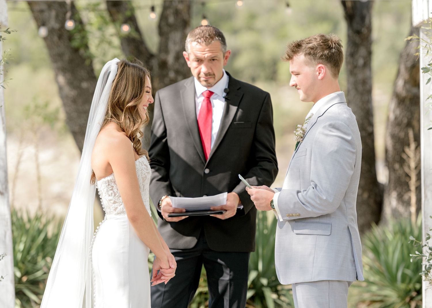 A wedding ceremony takes place outdoors with an officiant in a red tie conducting the service under trees.