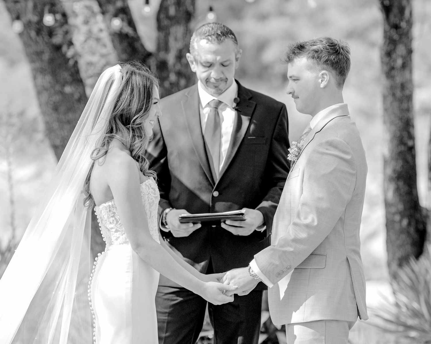 A black and white photo captures an intimate wedding ceremony moment under trees.