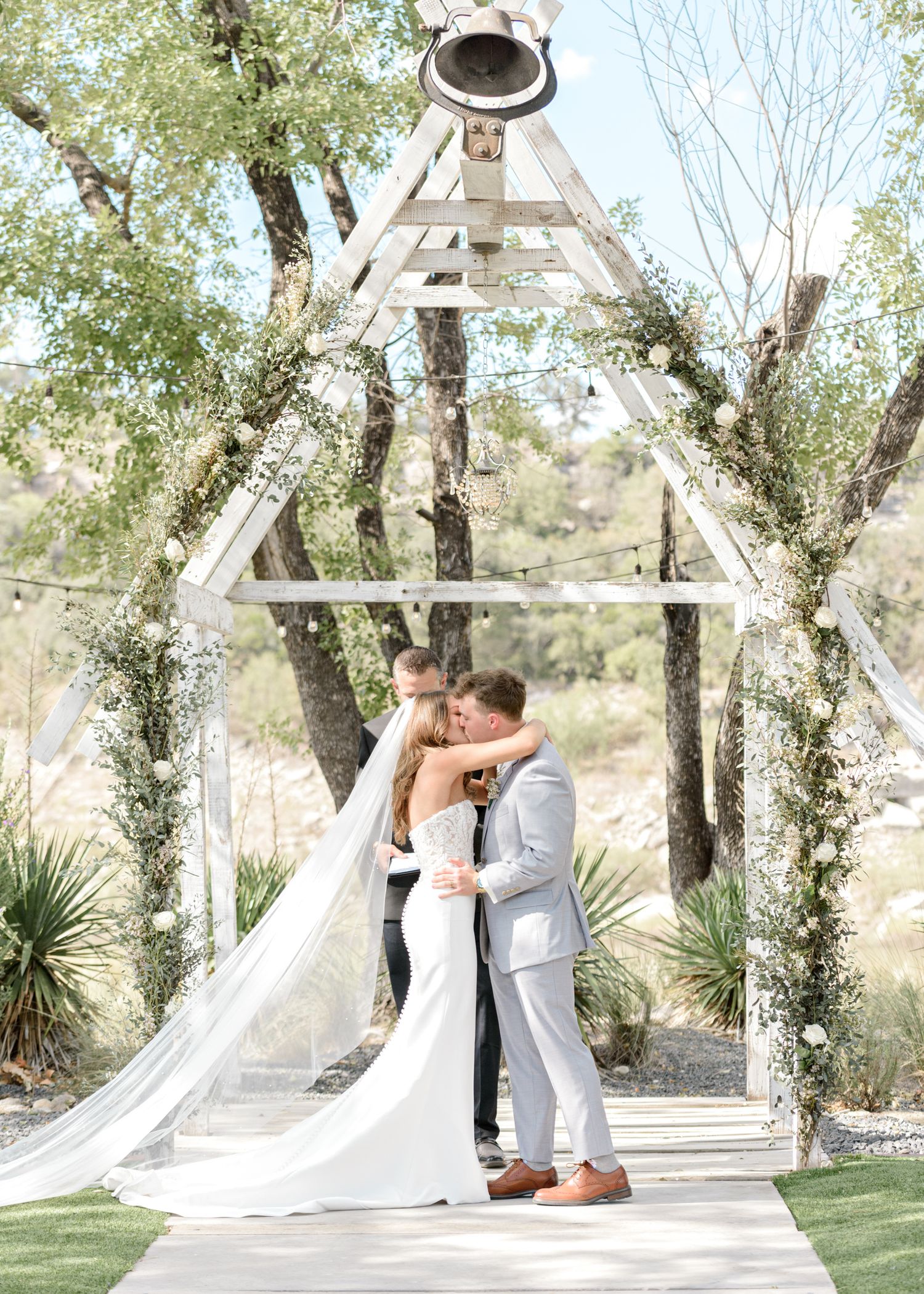 A romantic first kiss as newlyweds under a white wooden triangle arbor decorated with greenery.