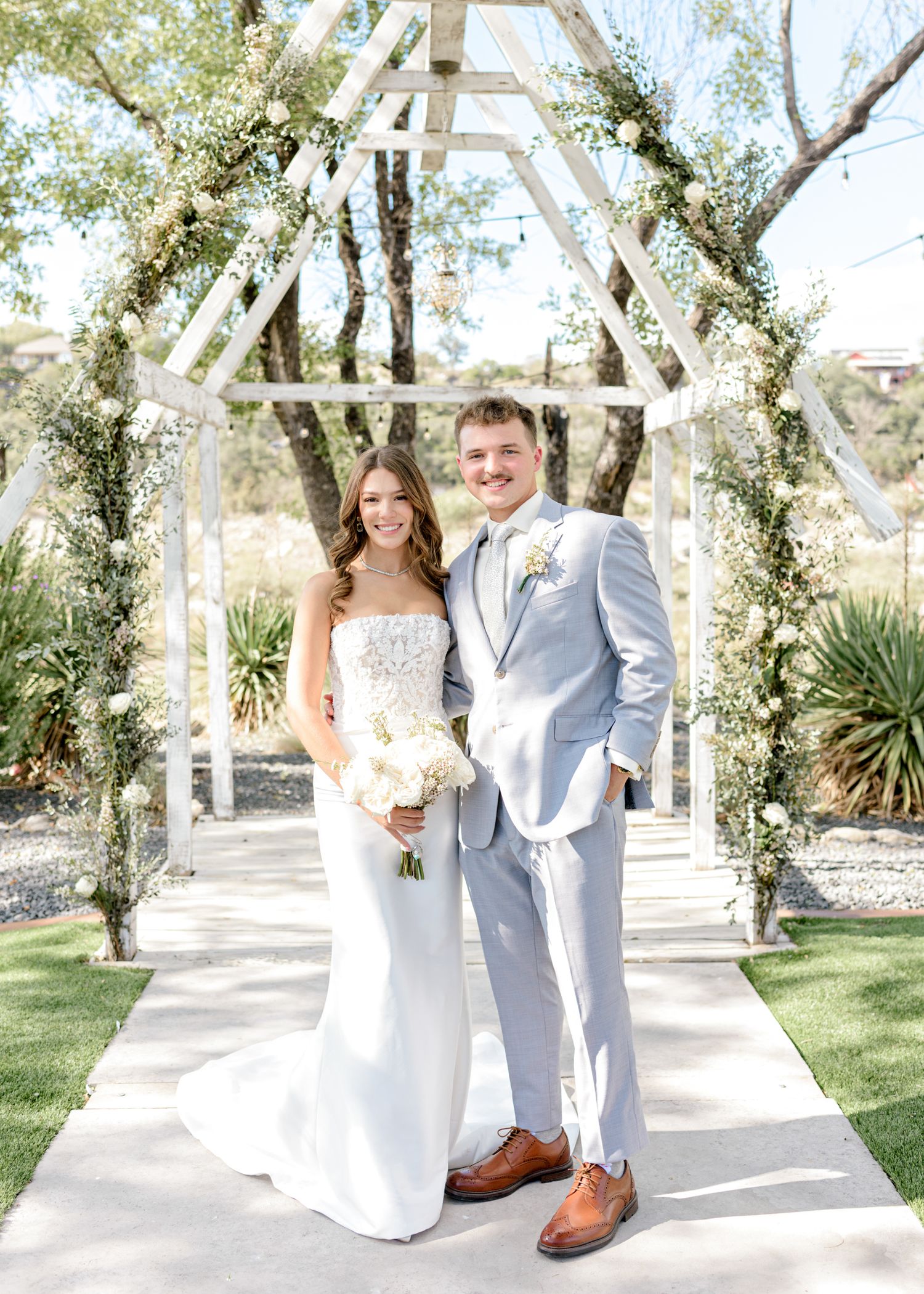 A couple poses under a white wooden triangle arch decorated with floral arrangements at their outdoor wedding.
