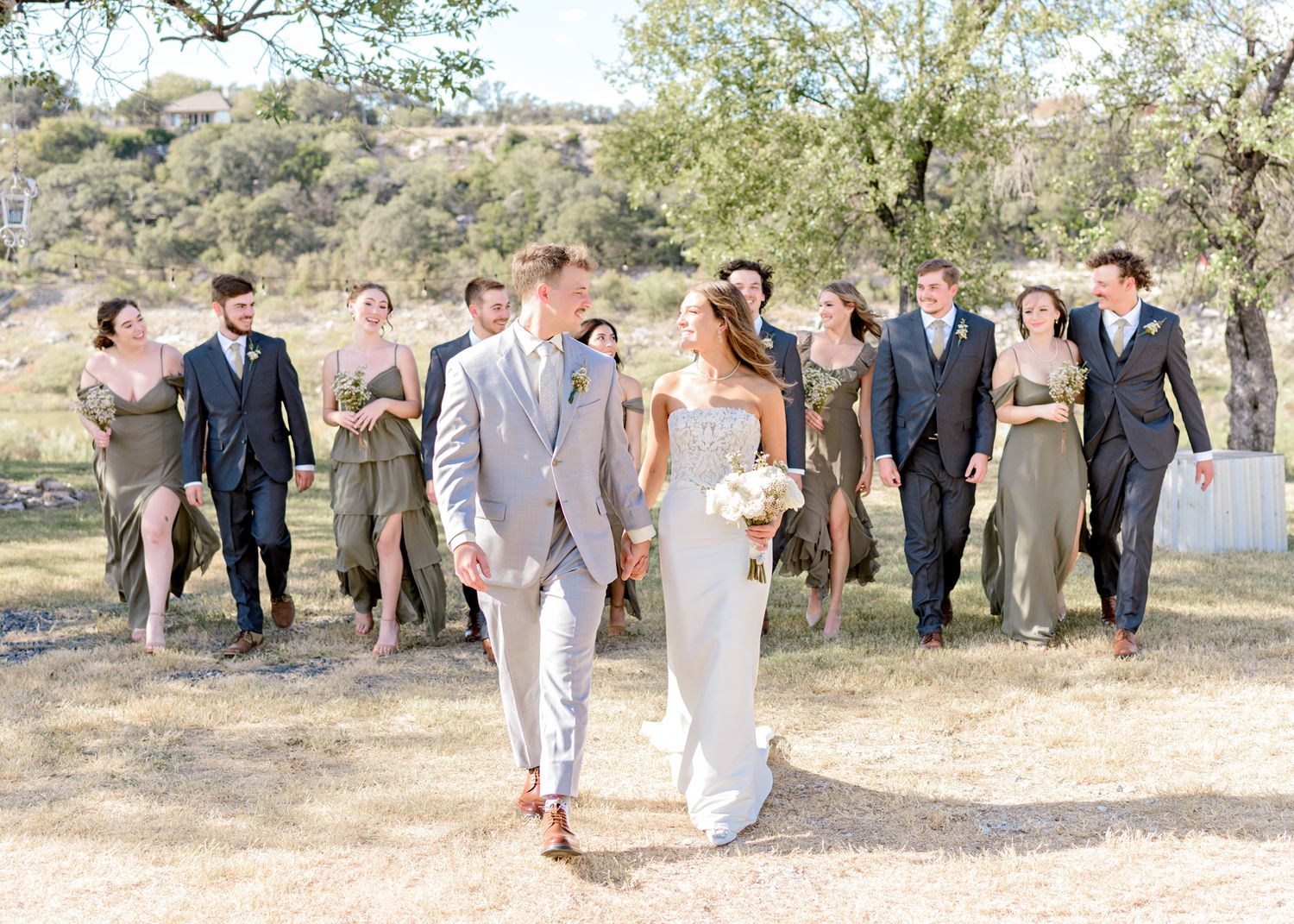 Wedding party walks joyfully through an outdoor venue with rolling hills and trees in the background.