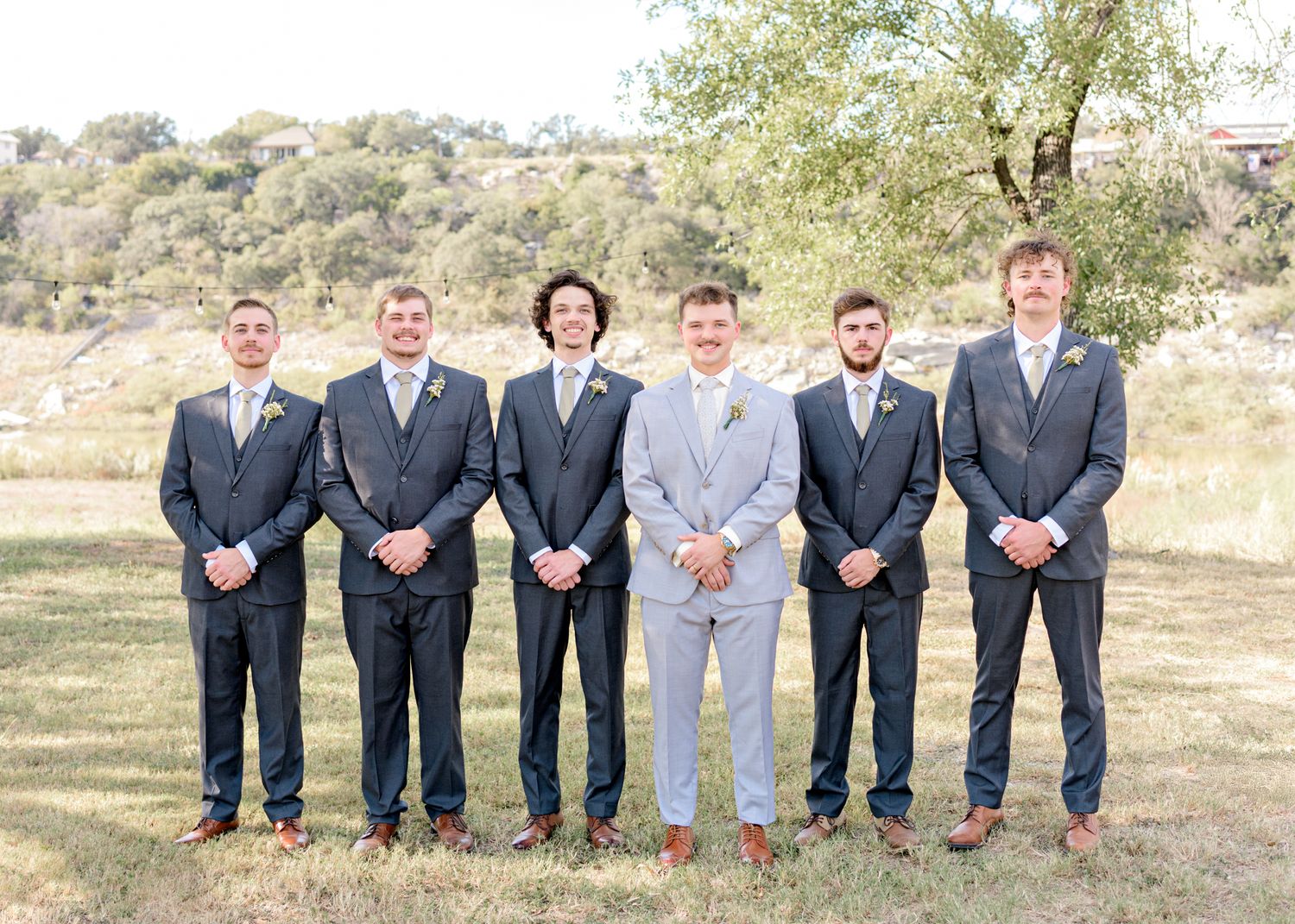 Groomsmen in dark suits with one in light gray pose formally outdoors.