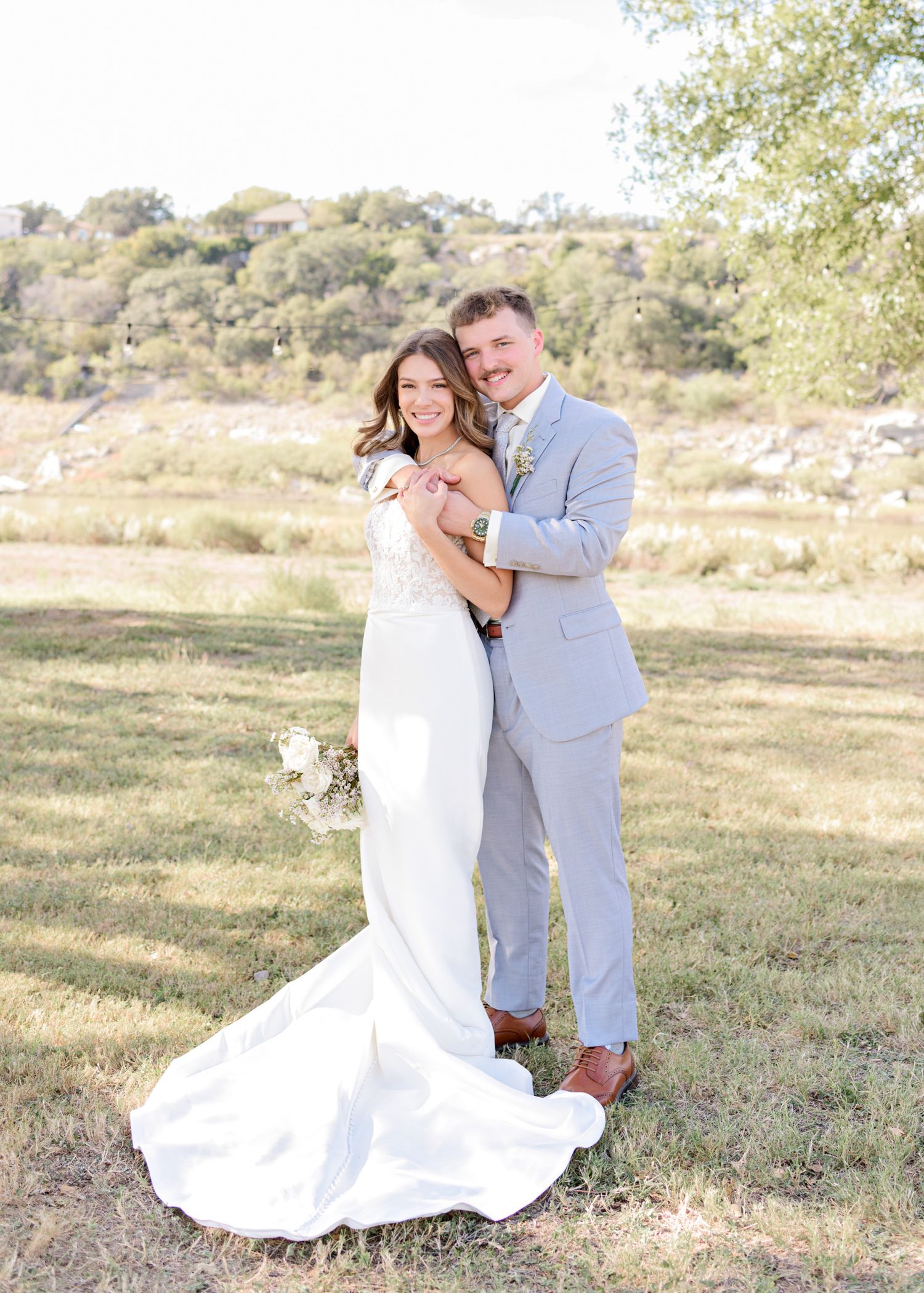 Couple in wedding attire embraces outdoors with rolling hills and trees in background.