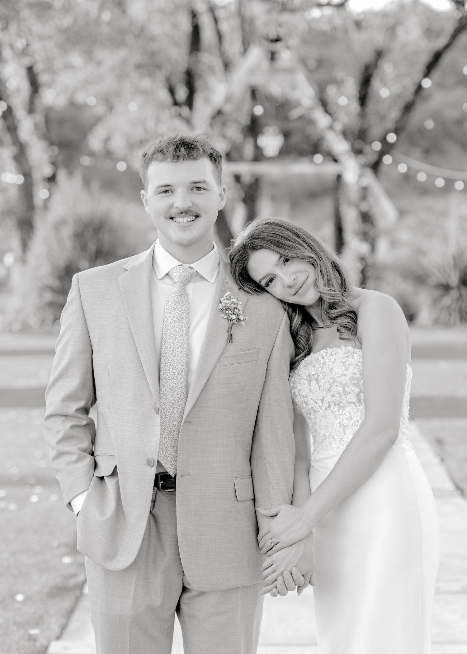 Black and white portrait of newlyweds in formal attire with string lights in background.