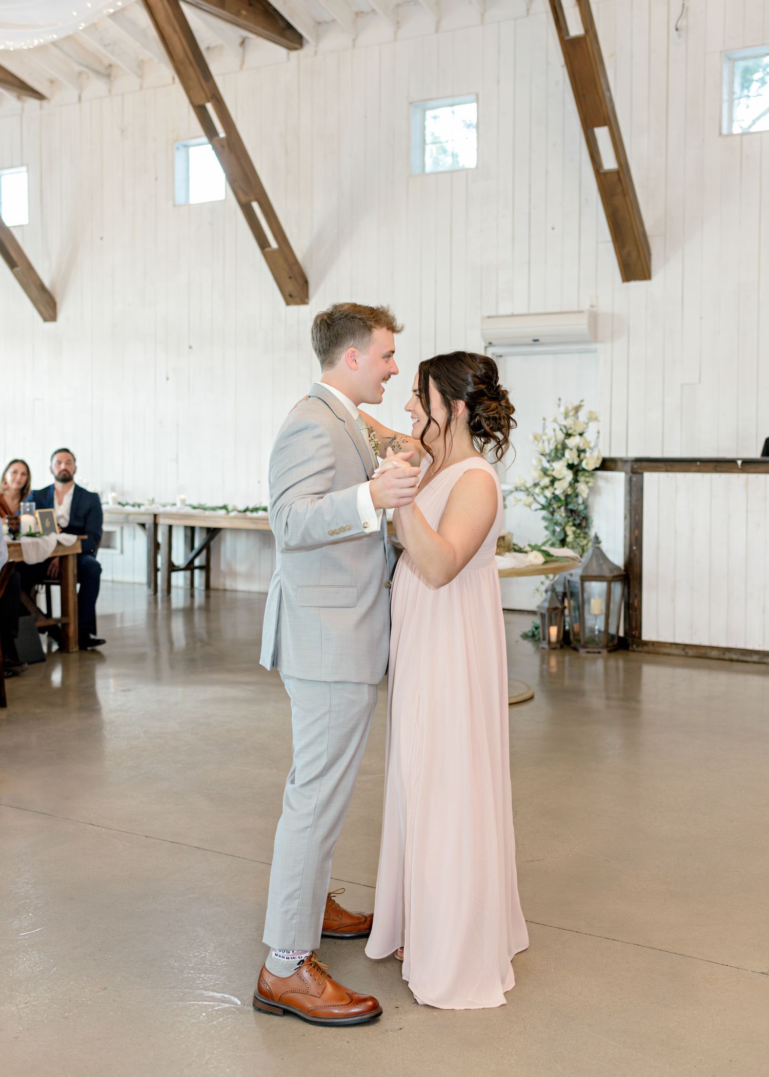 Couple dances together in bright barn venue while wedding guests look on from tables.