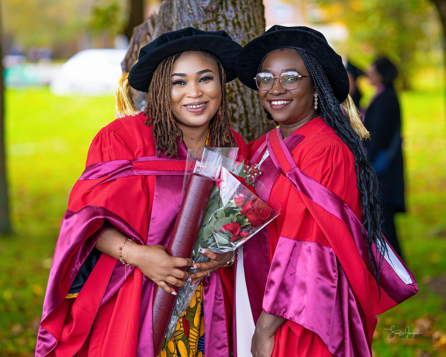 Two graduates in bright red academic regalia holding flowers and smiling during graduation ceremony outdoors.
