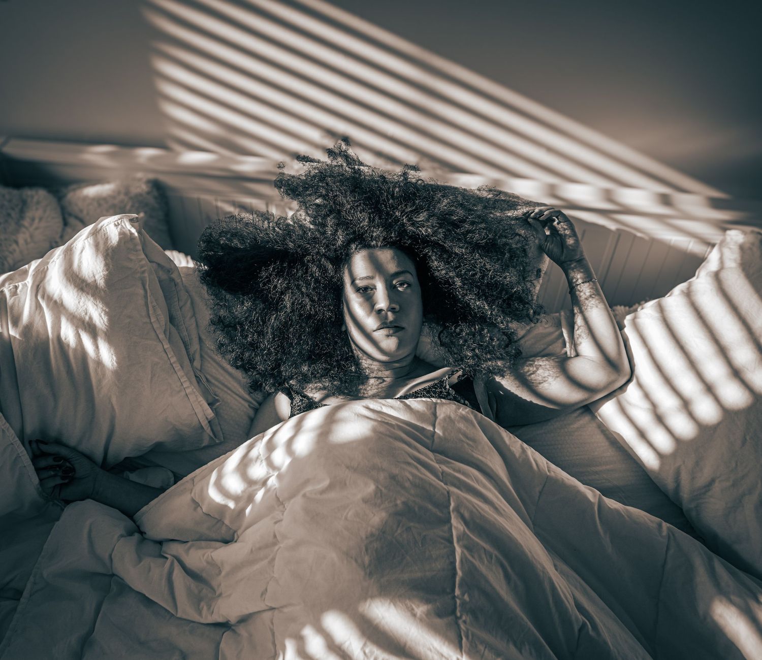 Black and white portrait of person with curly hair lying in bed with dramatic window shadows.