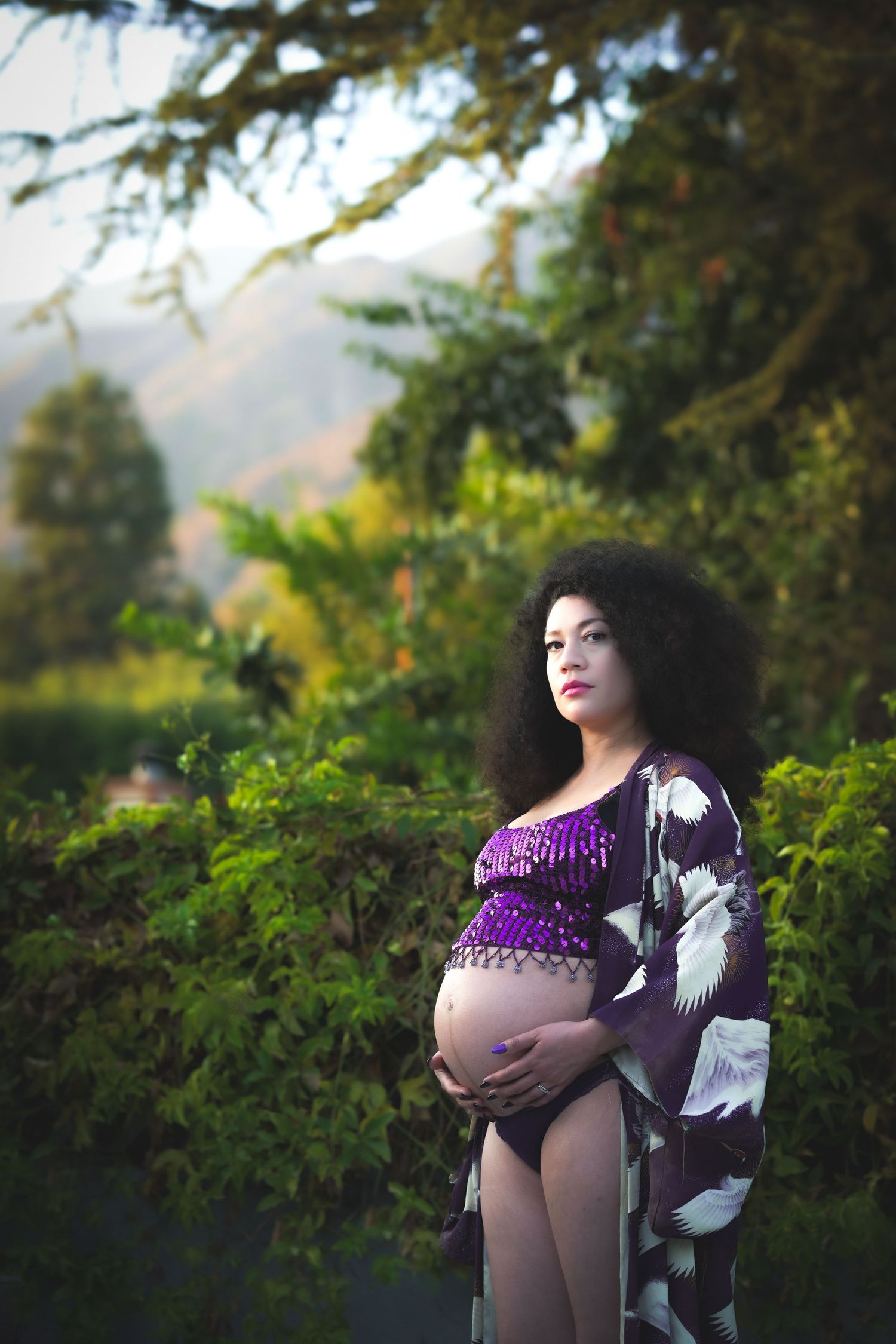 Outdoor maternity portrait with woman in purple dress amid lush garden scenery.