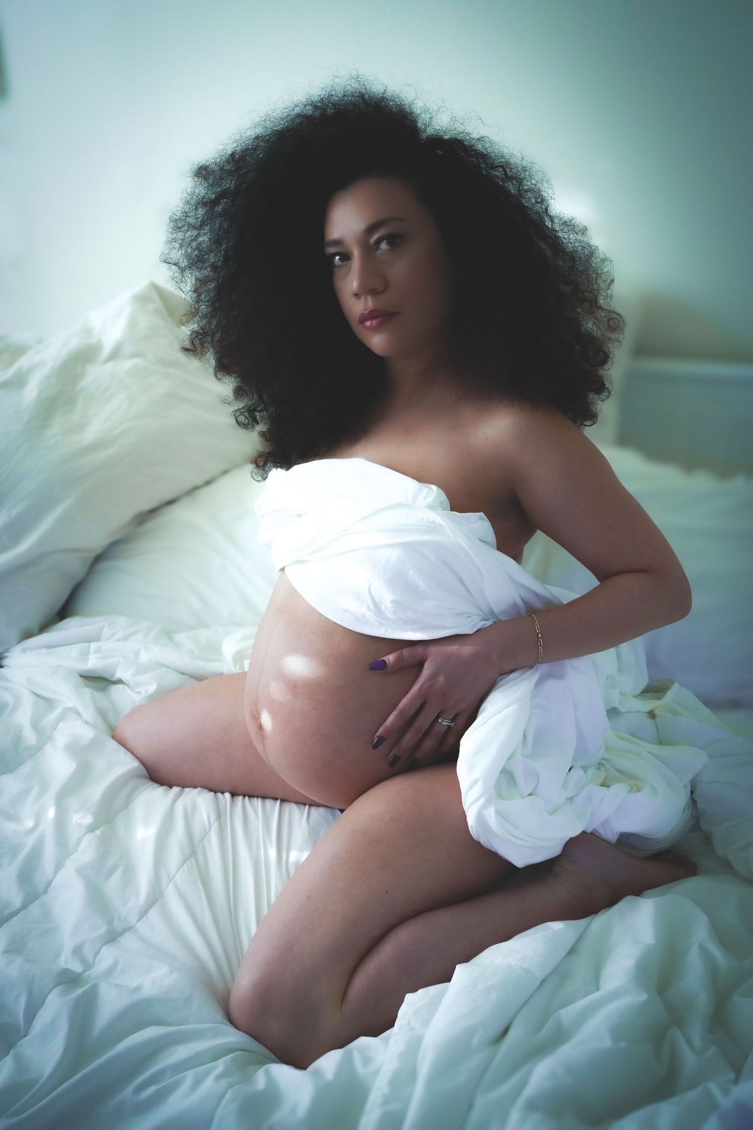 Artistic maternity photo of woman with curly hair draped in white sheets on bed.