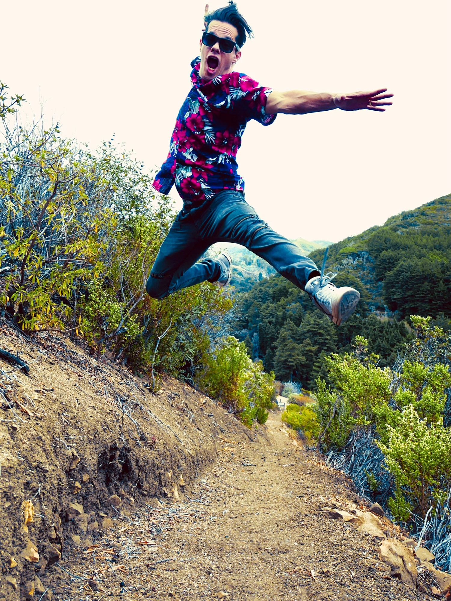 Energetic figure in colorful shirt leaps high on a hiking trail surrounded by green hills.