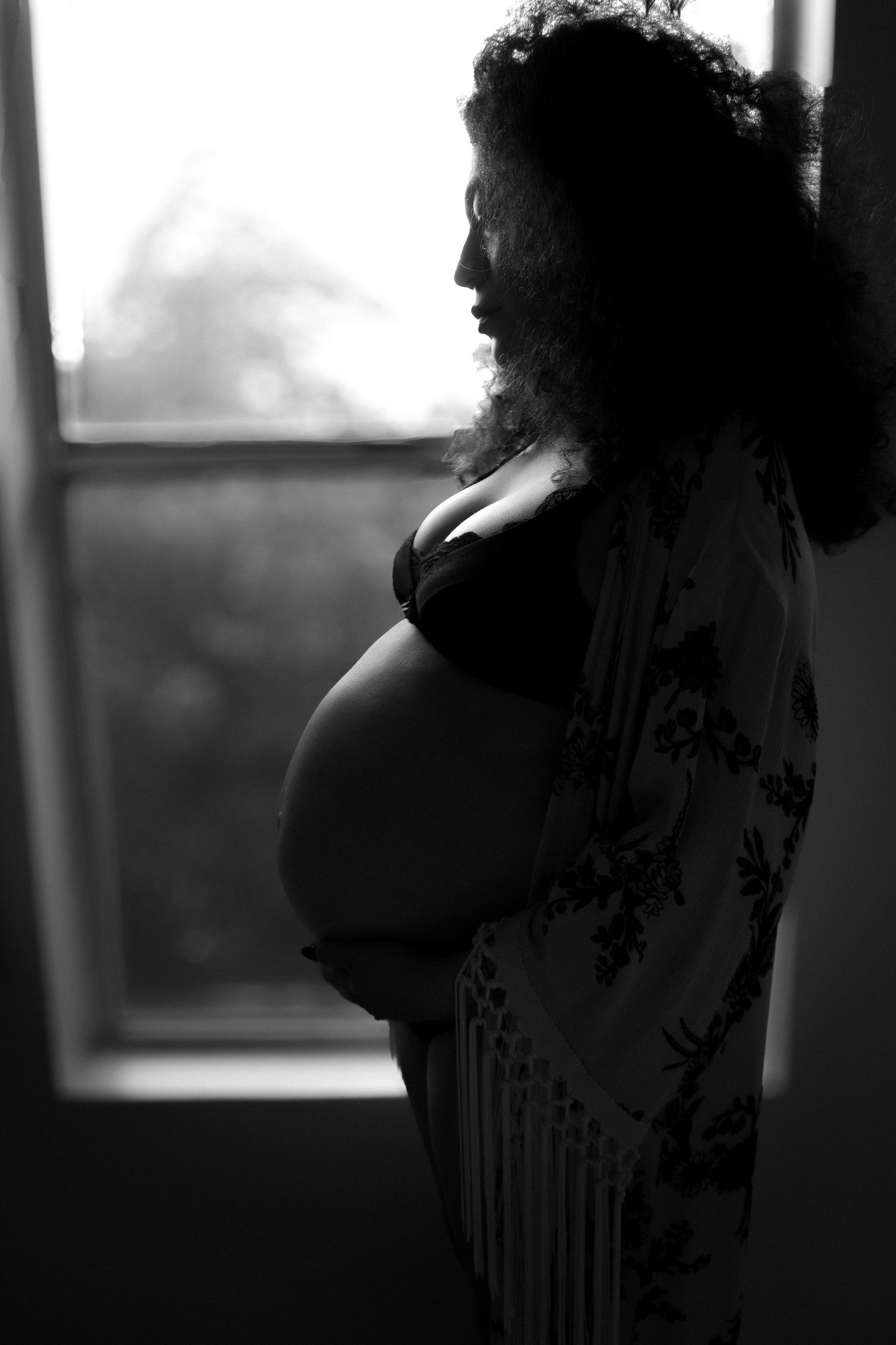 Black and white silhouette of an expectant mother in floral dress standing by window.