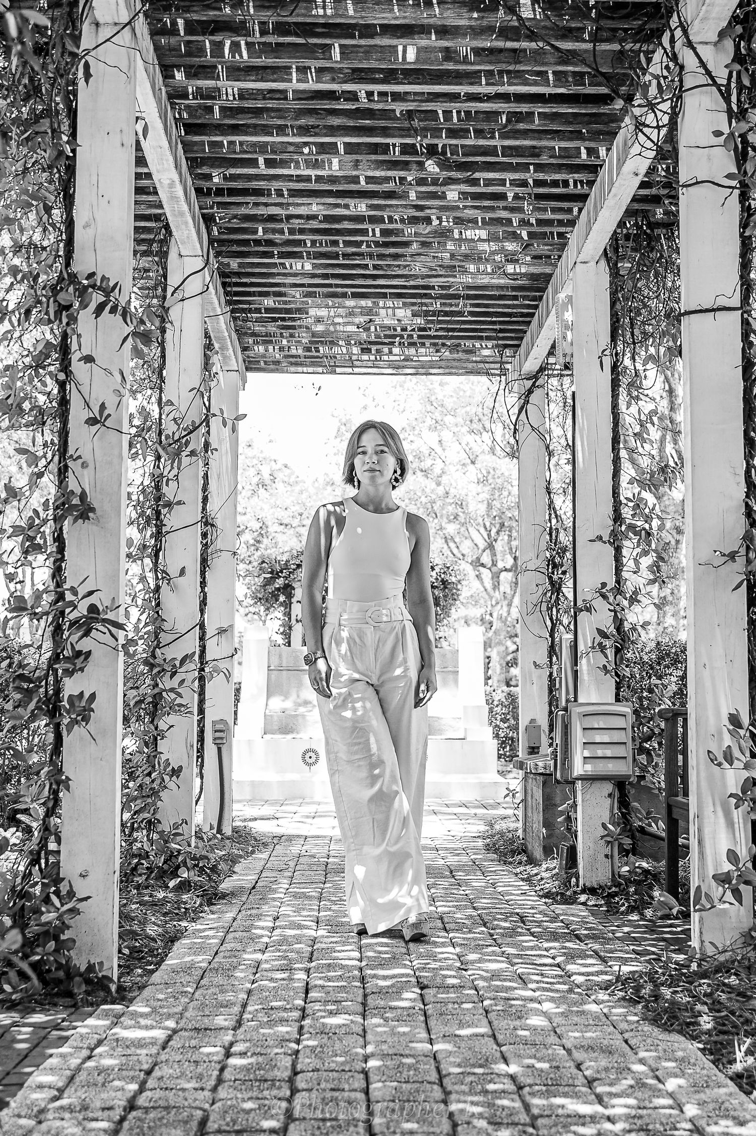 Black and white photo of an person standing in a brick archway covered with climbing vines at a garden.